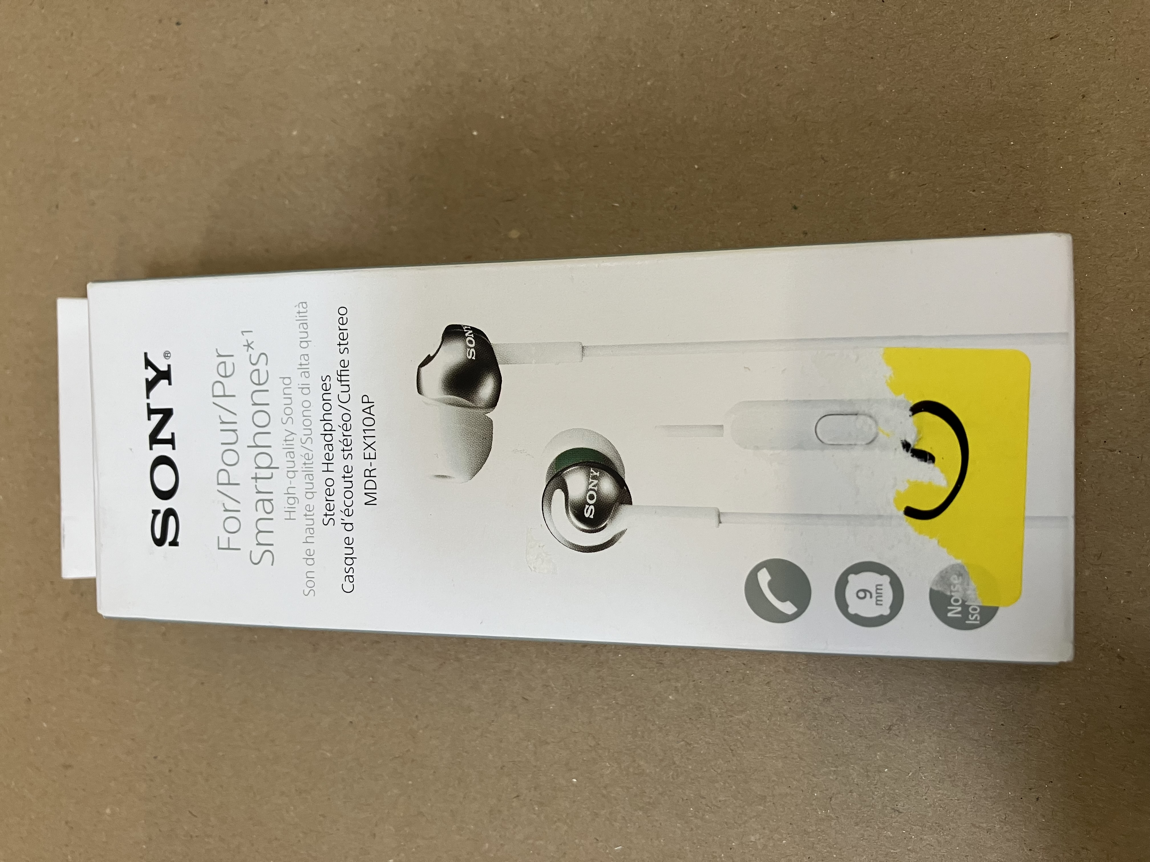 Niebieskie pudełko ze słuchawkami. Słuchawki w środku. Pudełko leży na brązowej powierzchni. Biały tekst i logo.