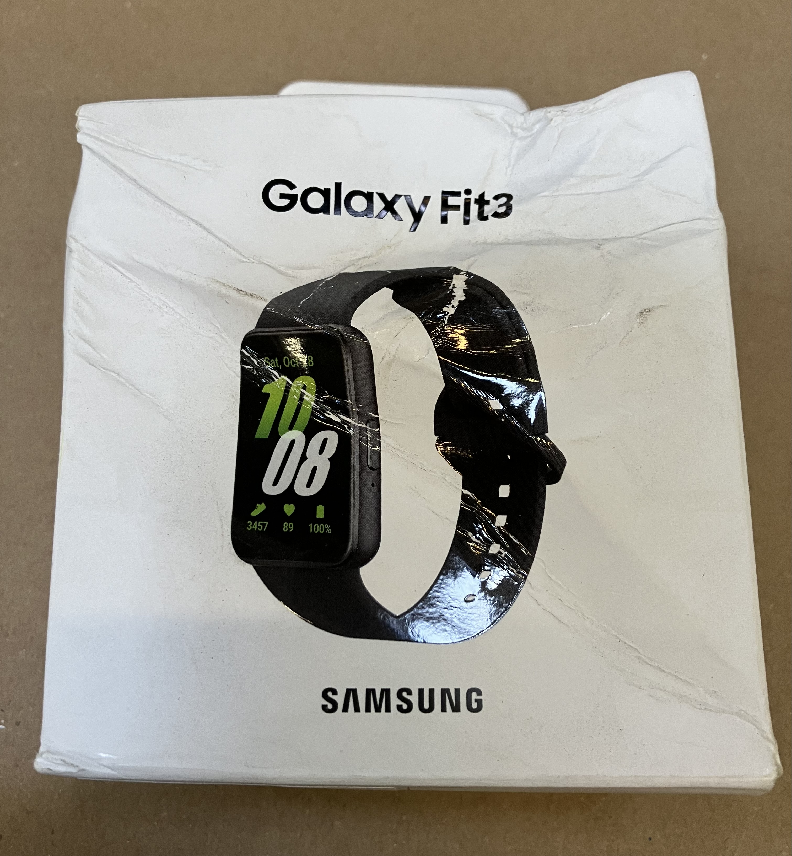 Smartwatch Galaxy Fit3 w białym pudełku, z czarnym paskiem i ekranem wyświetlającym czas.
