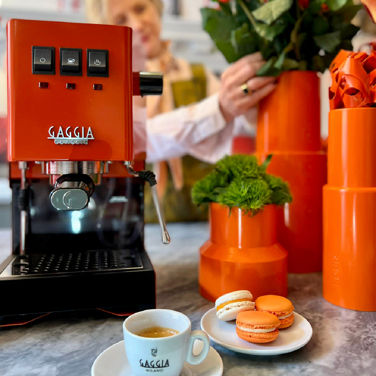 Eine orangefarbene Espressomaschine, orangefarbene Vasen und ein Teller mit Makronen. Eine Person im Hintergrund.