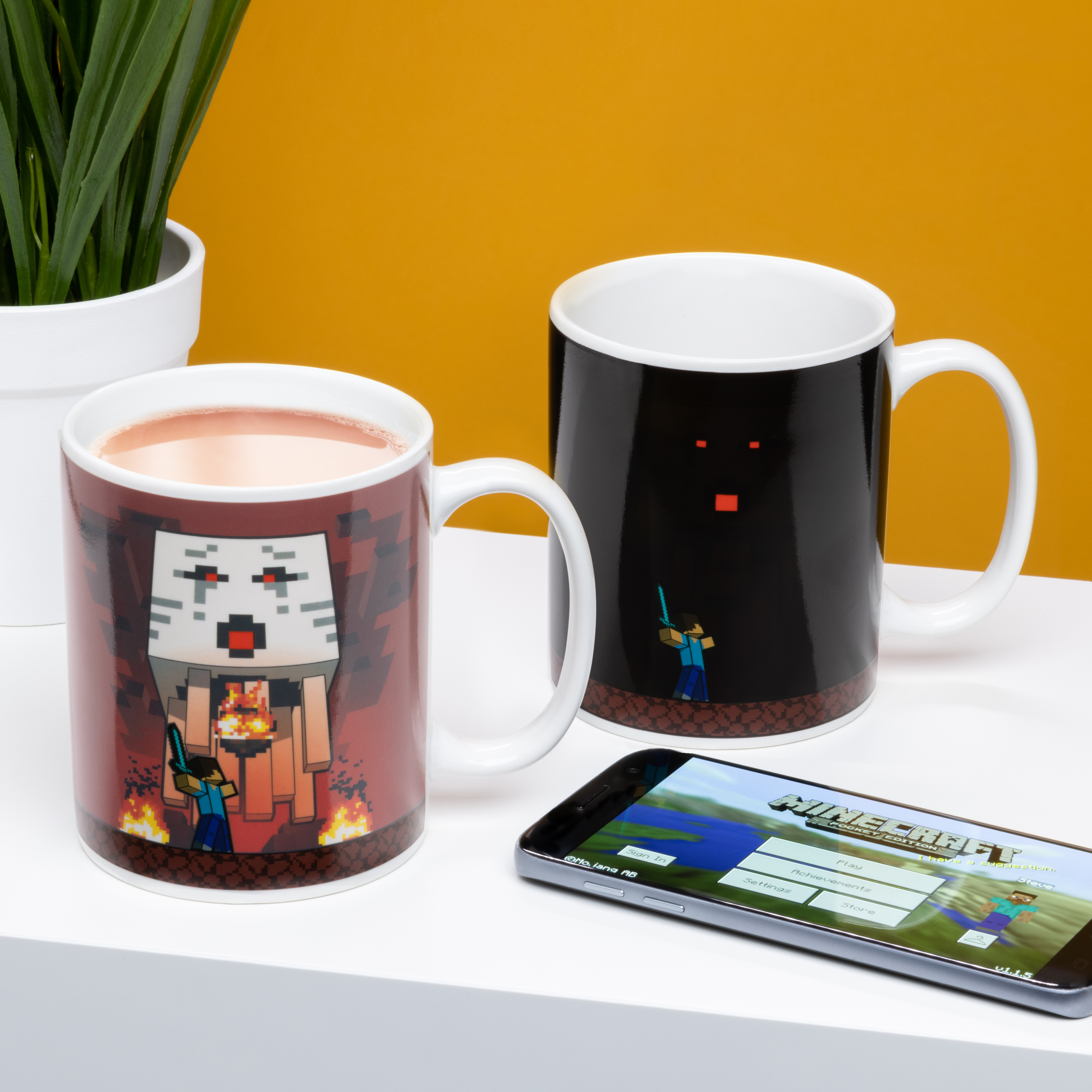 Zwei Minecraft-Tassen, eine mit Pixelkunst, und ein Telefon mit Spielbildschirm.