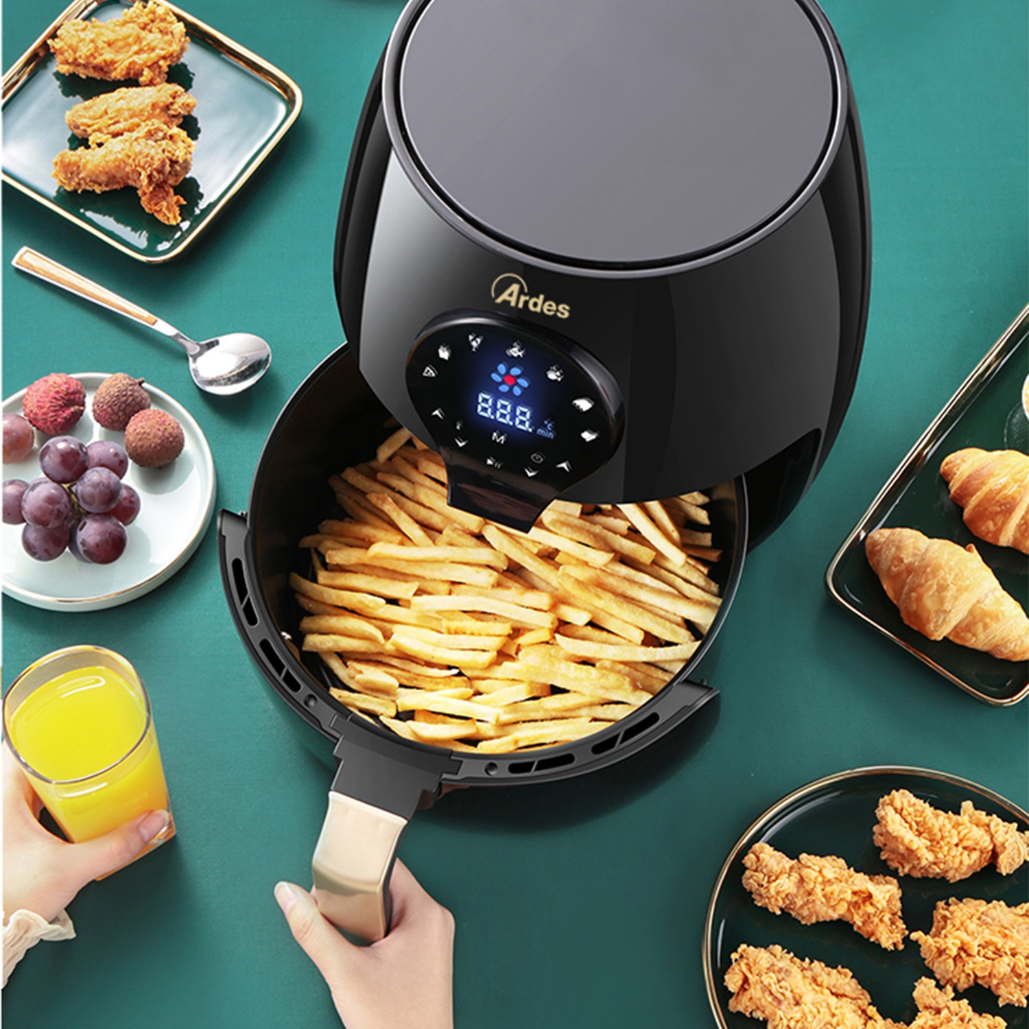 Una friggitrice ad aria piena di patatine fritte. Ci sono piatti con cibo intorno e una persona tiene la maniglia.
