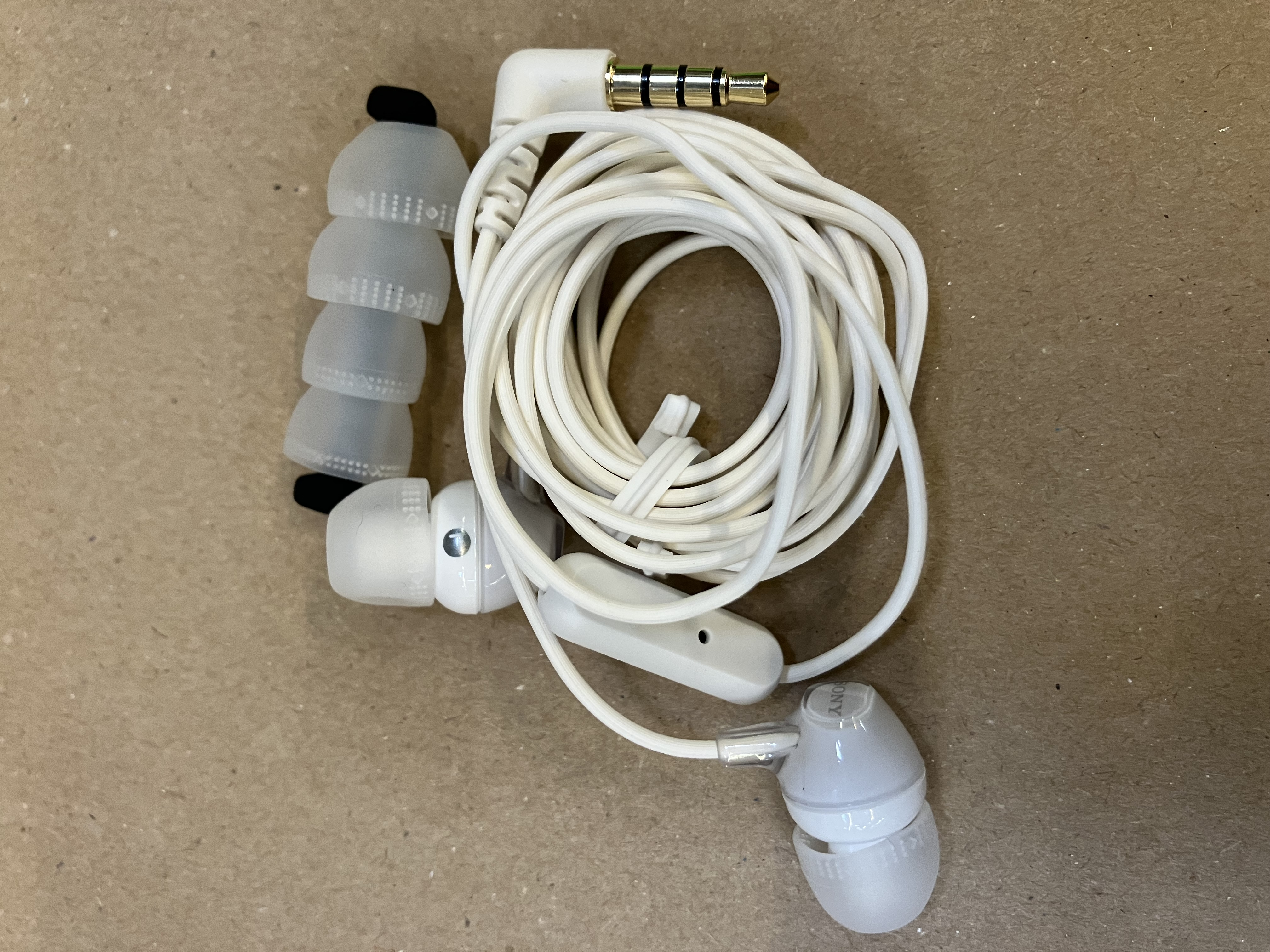 Białe słuchawki z dodatkowymi wkładkami dousznymi i przewodem. Prezentowane na brązowej powierzchni.