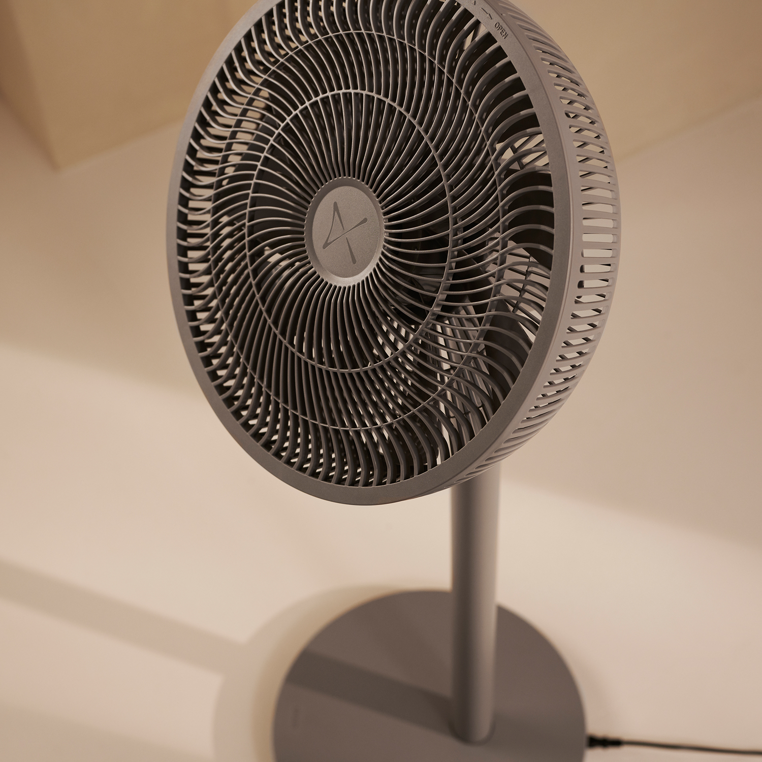 Grijze ventilator op een standaard, geplaatst in een kamer met een witte muur.