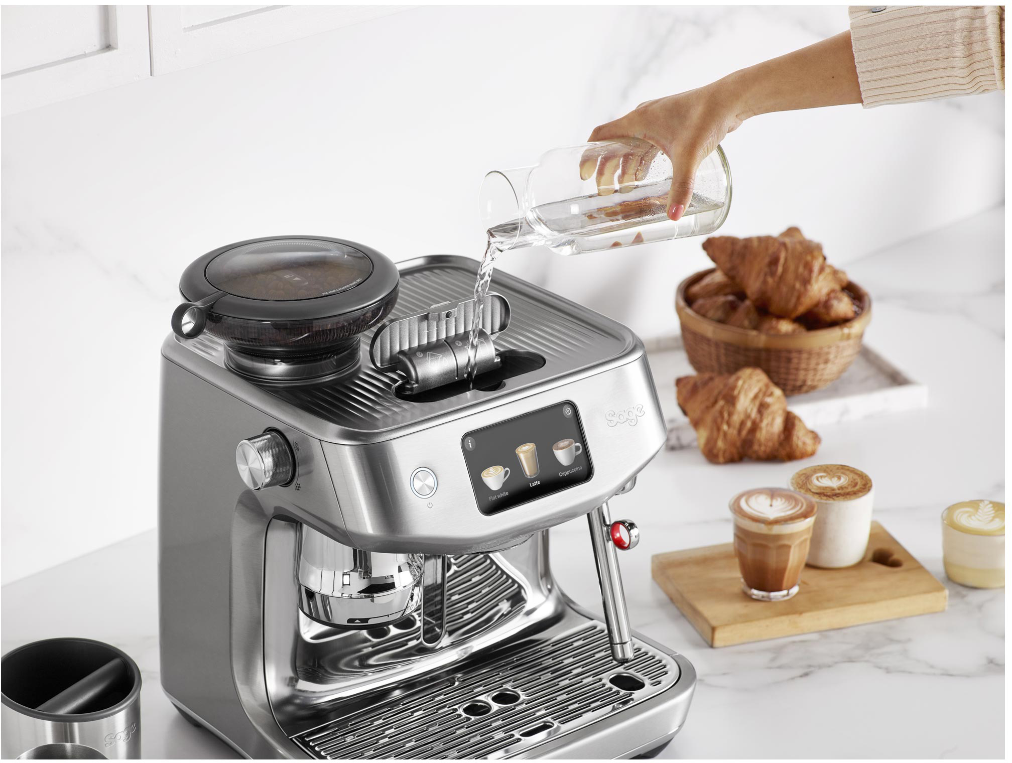 Egy személy vizet önt egy ezüst eszpresszógépbe, croissantok és kávéscsészék az oldalán.