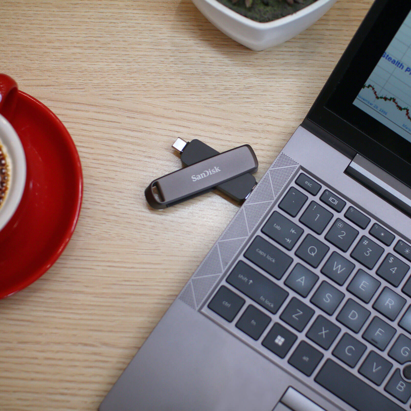 Laptop mit USB-C-Laufwerk auf dem Holztisch. Eine rote Tasse und eine Pflanze sind ebenfalls sichtbar.