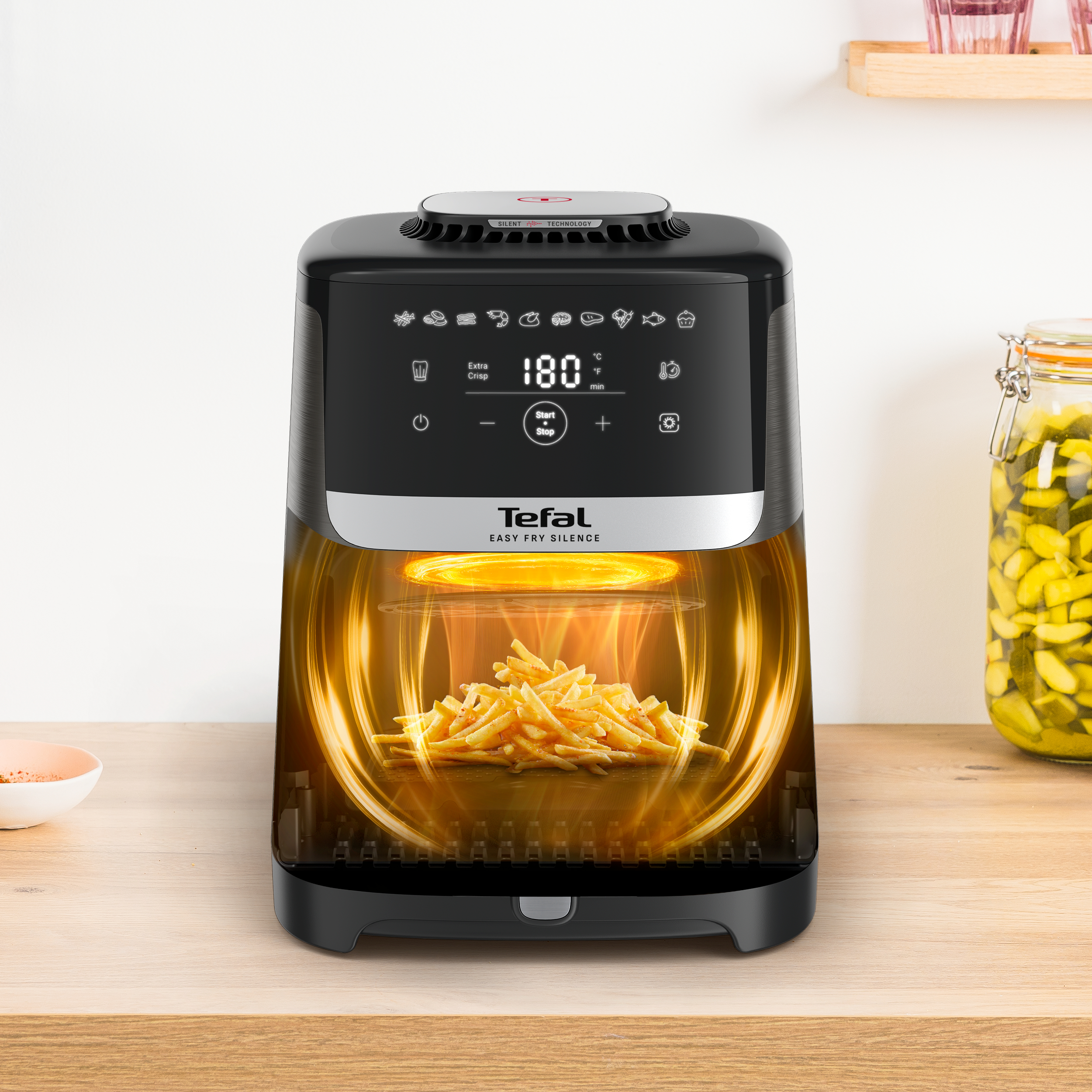 Tefal-Heißluftfritteuse mit Pommes darin. Schwarzer Körper, Digitalanzeige. Holzoberfläche, Glas im Hintergrund.