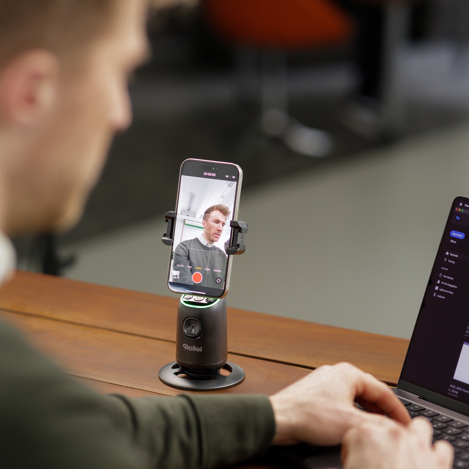 A man is recording a video with his phone on a stand, with a laptop next to it.