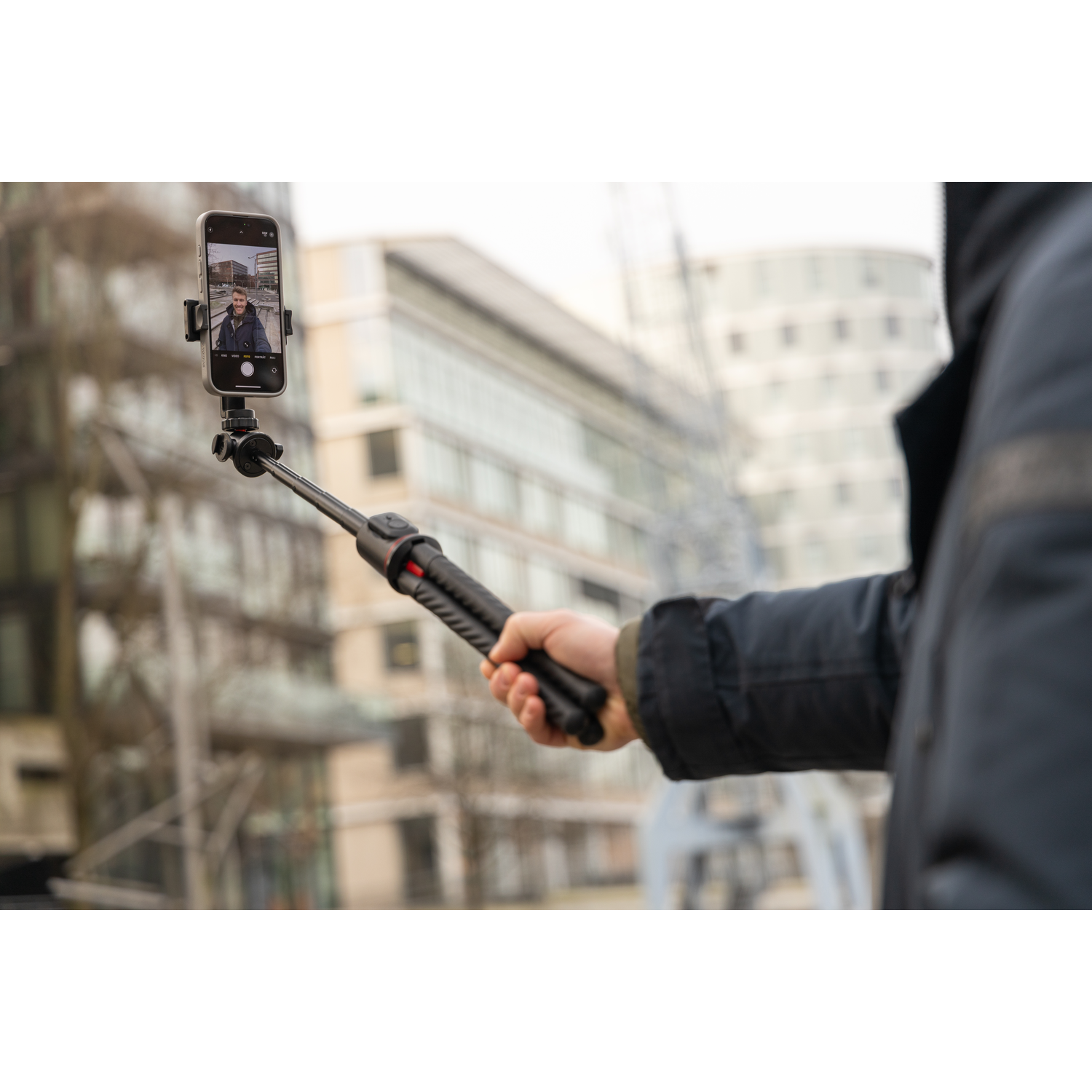 Person holding a selfie stick with a phone, taking a photo. Buildings in the background.
