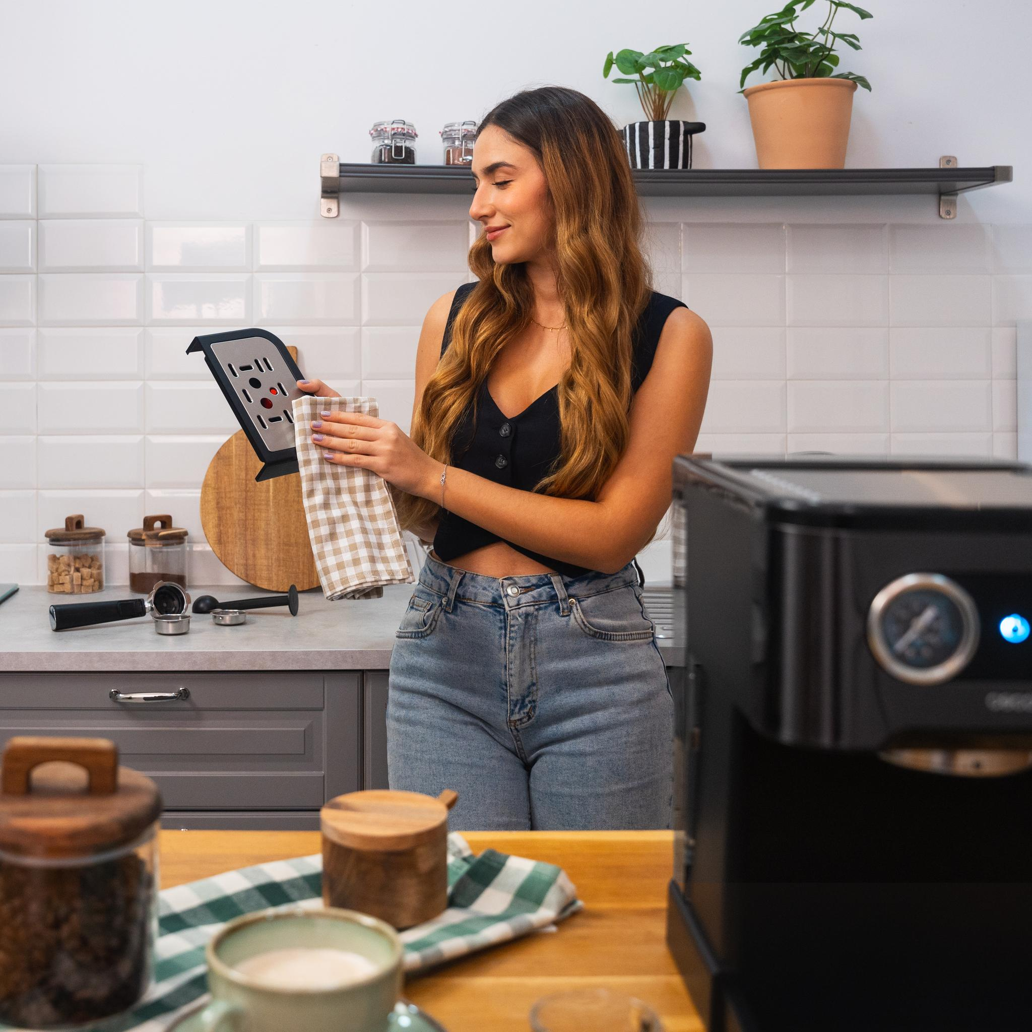 Una mujer en una cocina sosteniendo una toalla y una cafetera. Hay plantas en el fondo.