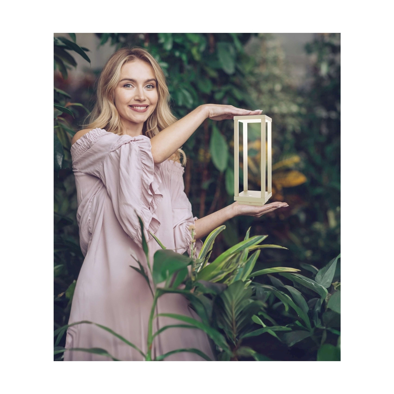 Donna che tiene in mano una lampada quadrata in giardino. Piante verdi. Sorridente.
