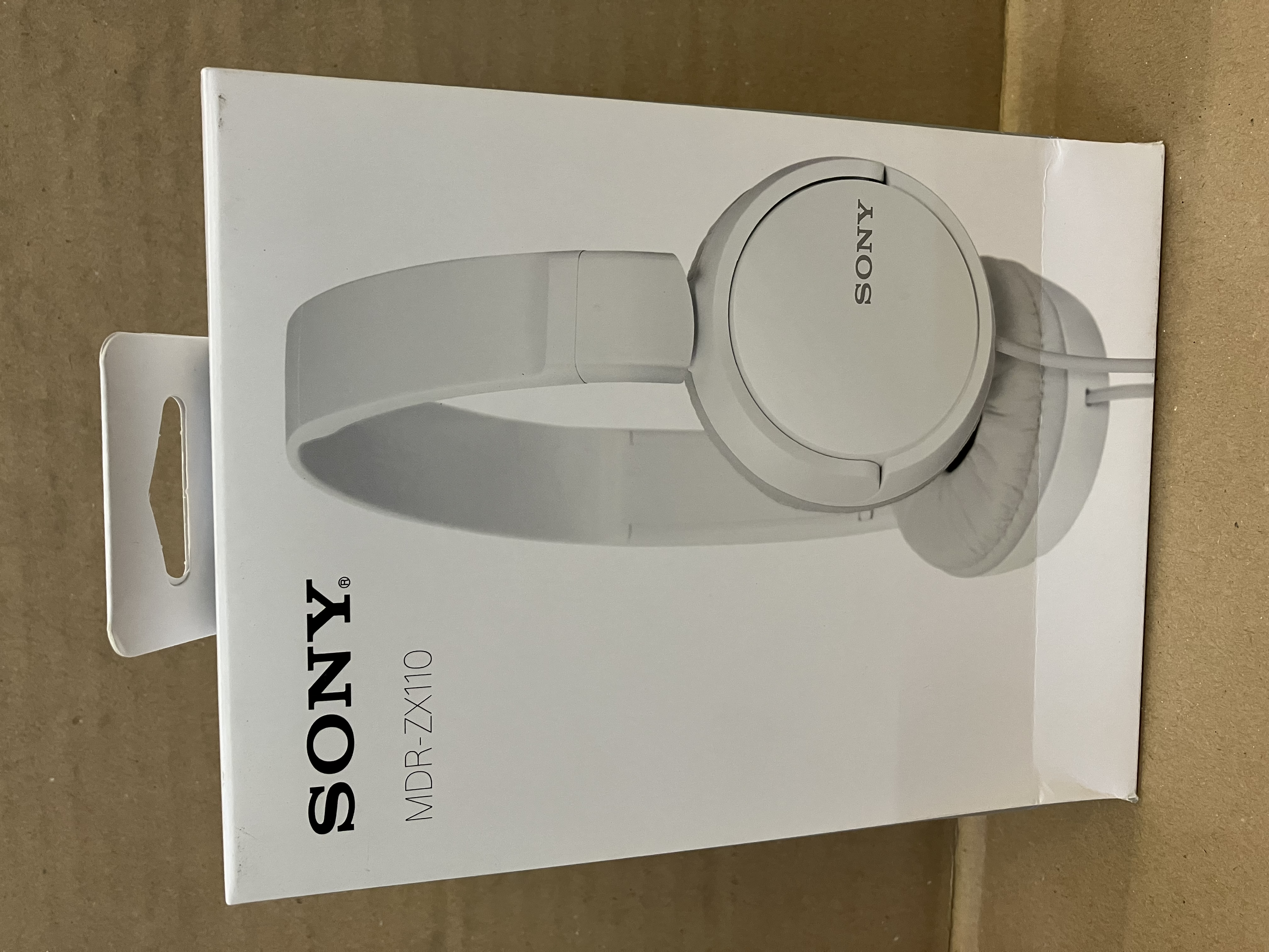 Białe pudełko ze słuchawkami SONY. Pudełko znajduje się na brązowej powierzchni.