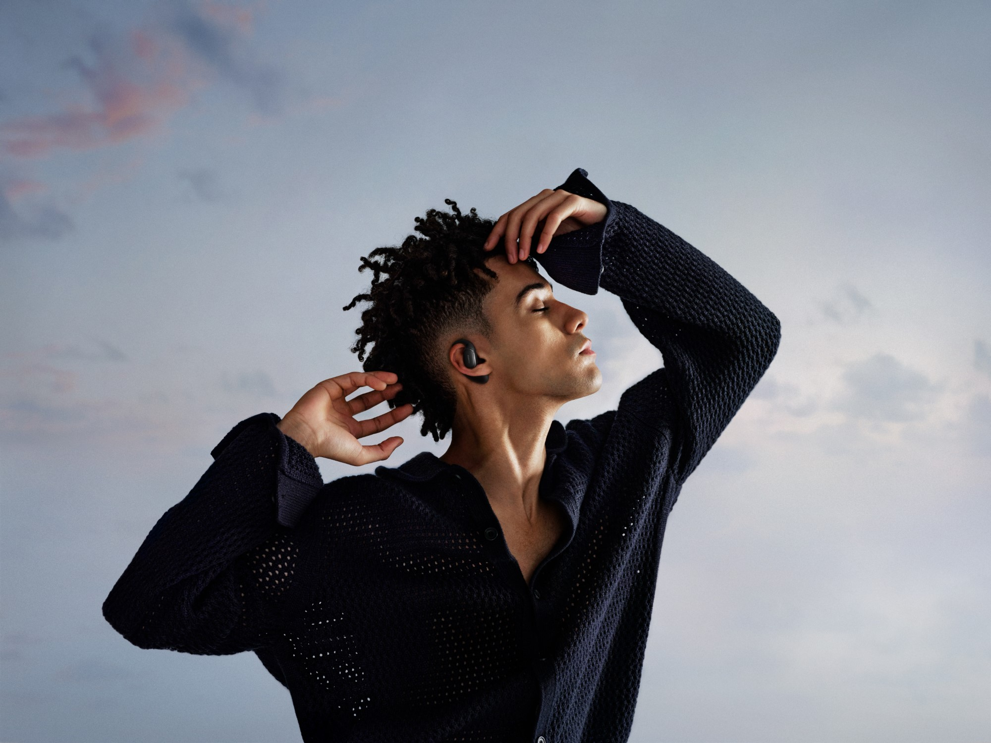 Młody mężczyzna z kręconymi włosami w ciemnym swetrze i słuchawkach bezprzewodowych, tło nieba.