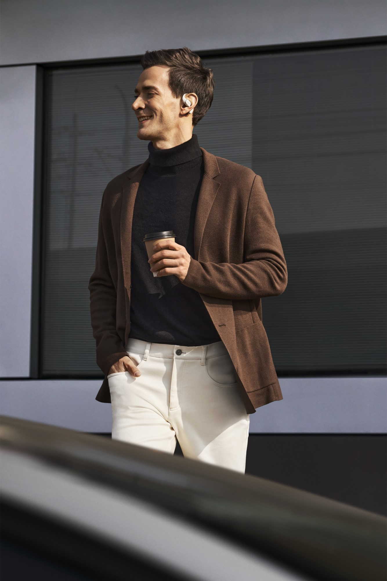 Mężczyzna idzie z kubkiem kawy i słuchawkami, uśmiechając się. Ma na sobie marynarkę, golf i białe spodnie.