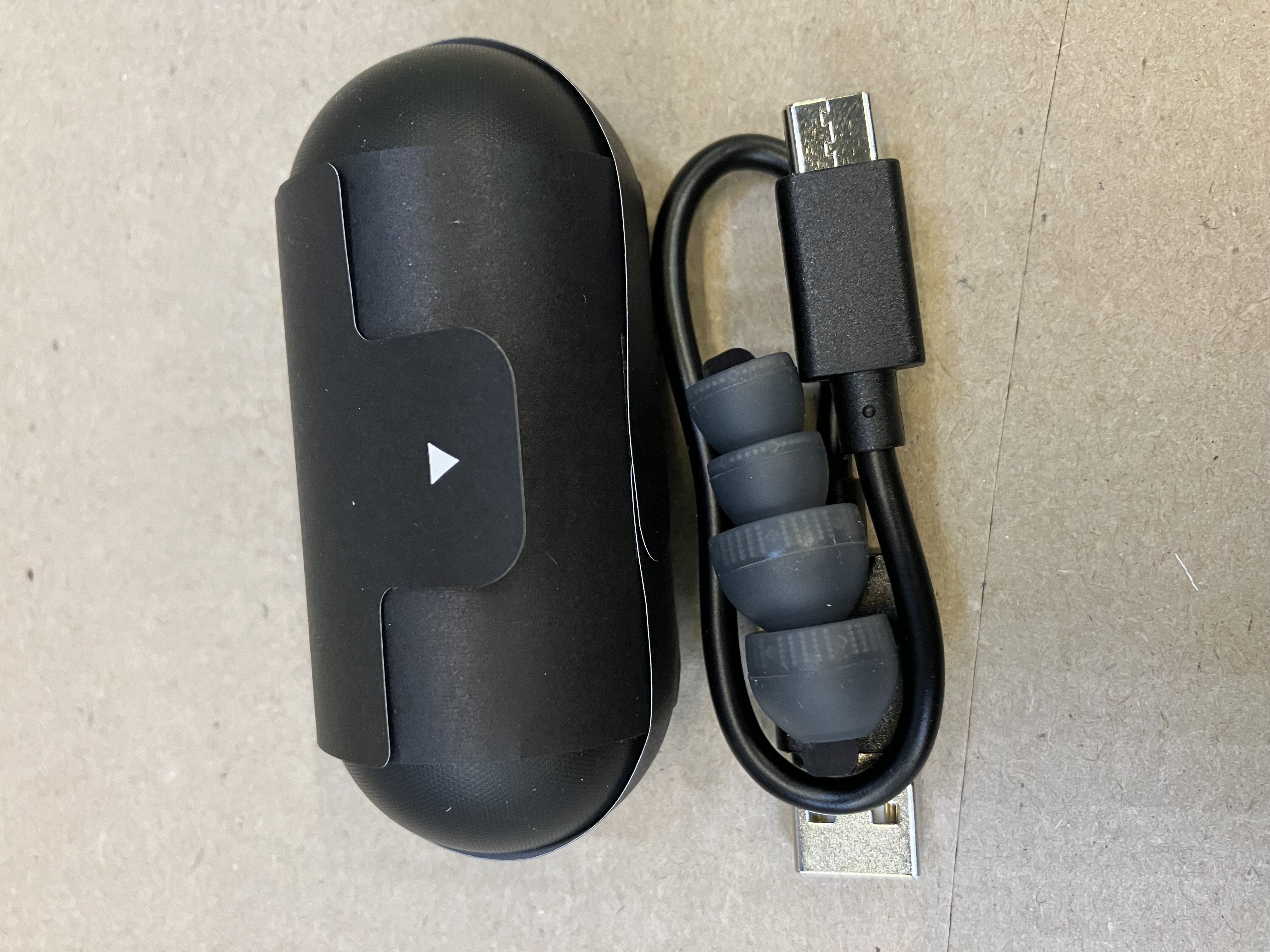 Czarny futerał na słuchawki z białą strzałką, szare wkładki i kabel USB-C. Na brązowej powierzchni.