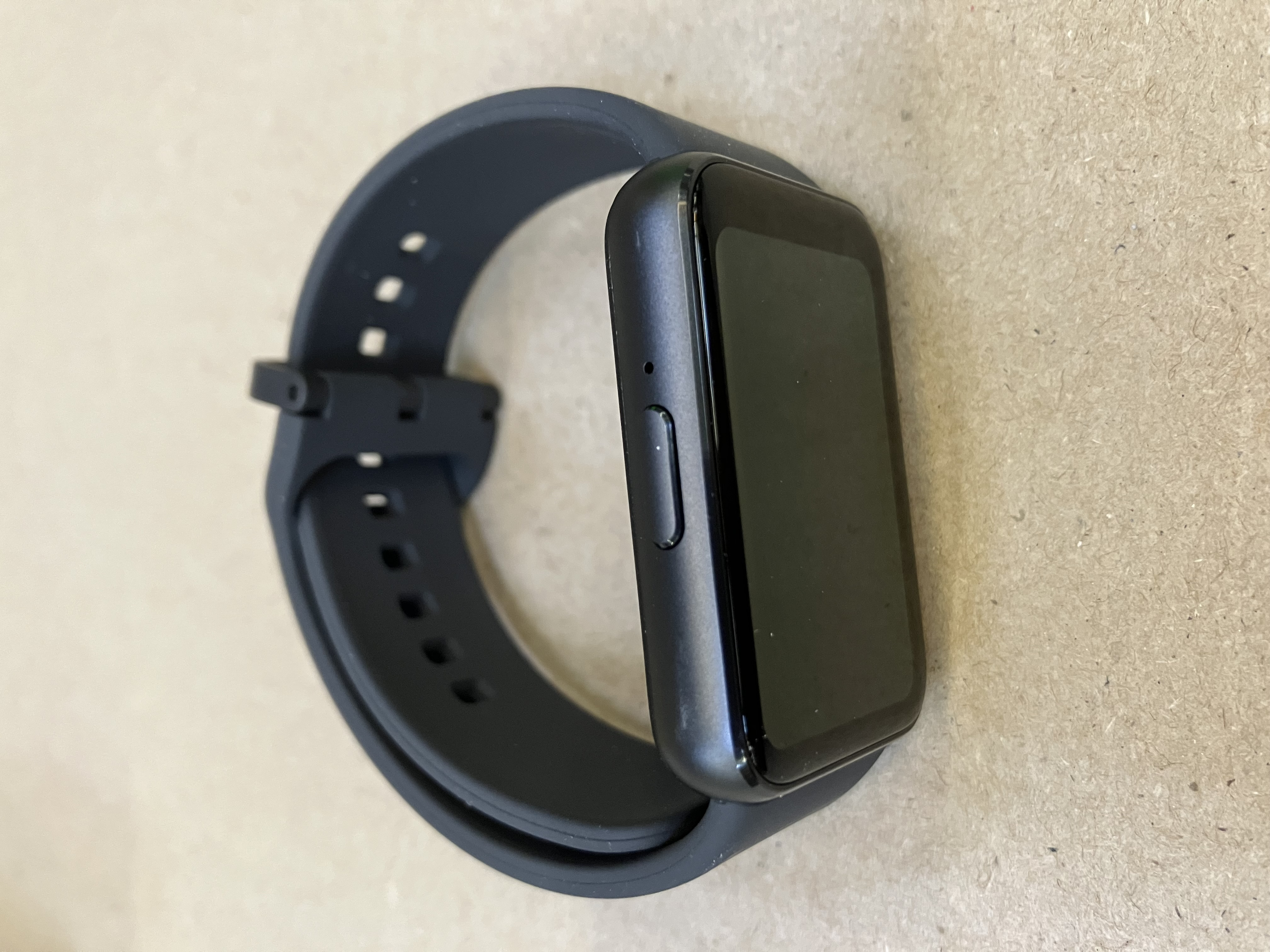 Czarny smartwatch z czarnym paskiem na drewnianej powierzchni. Widoczny przycisk boczny.