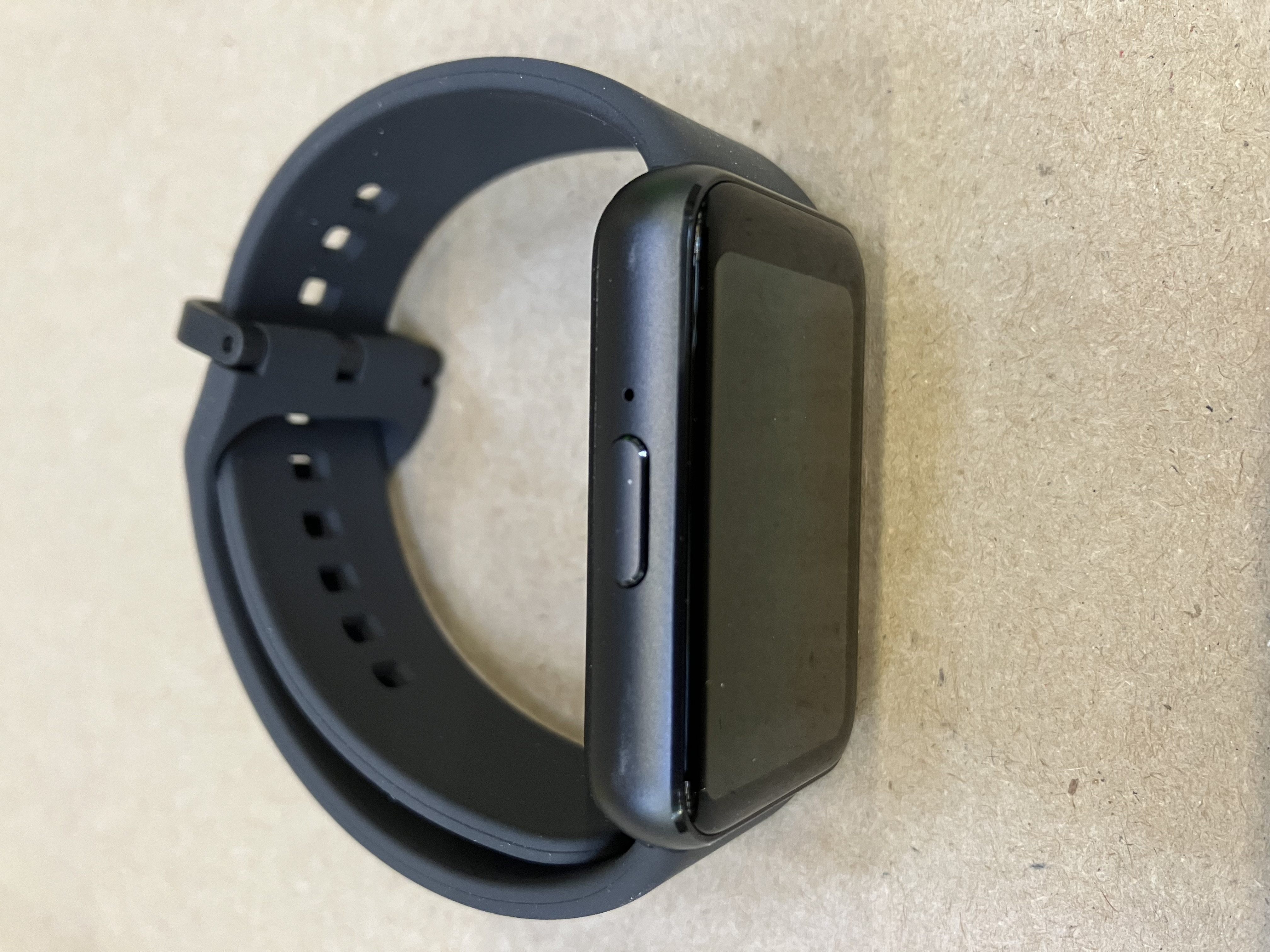 Ciemnoszary smartwatch z czarnym ekranem i czarnym paskiem. Zegarek leży na drewnianej powierzchni.