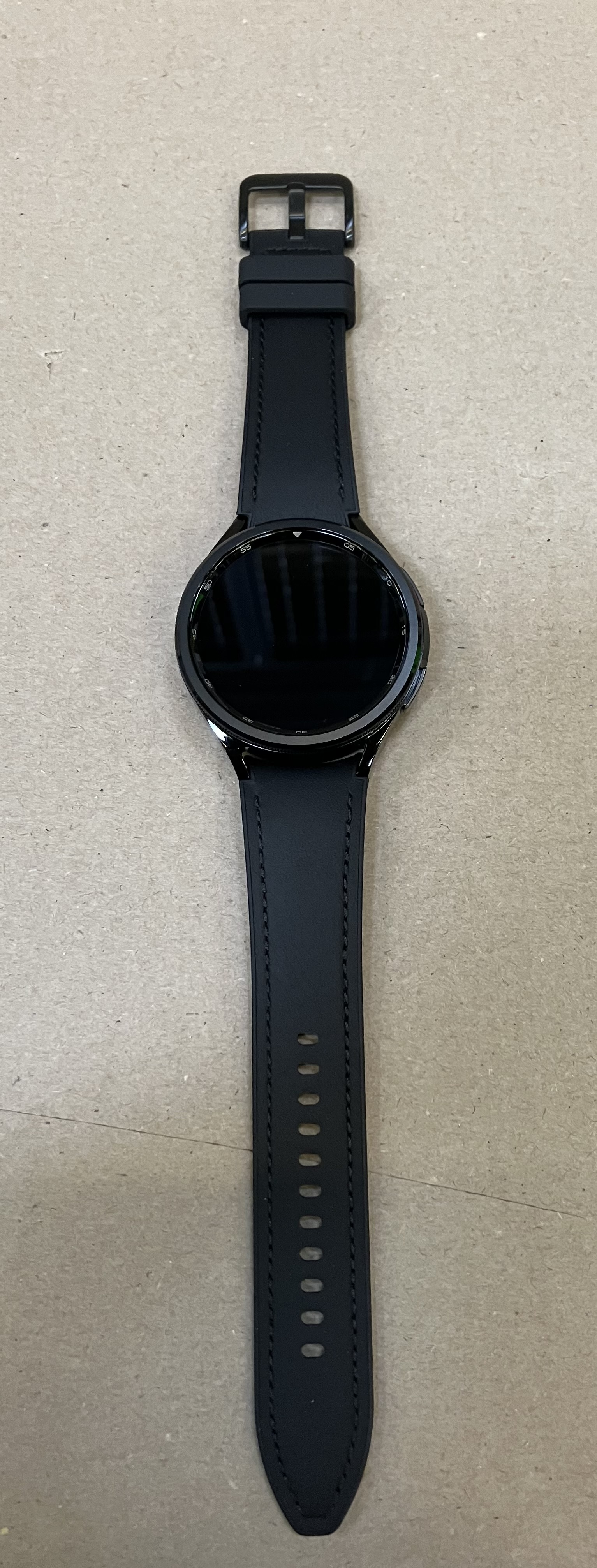 Czarny smartwatch z czarnym skórzanym paskiem na szarej powierzchni.