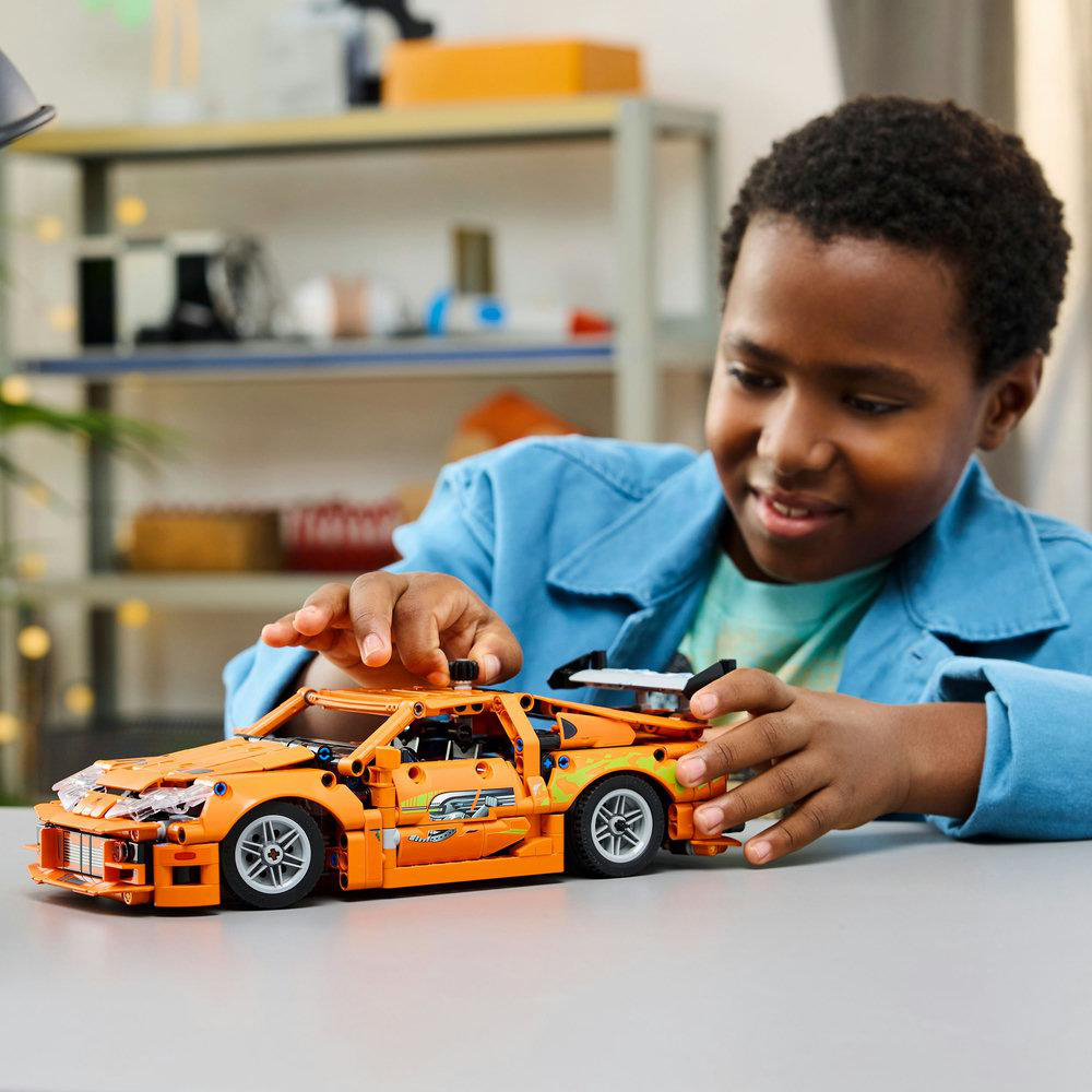 Ein Junge baut ein orangefarbenes Lego-Auto auf einem grauen Tisch, lächelnd.