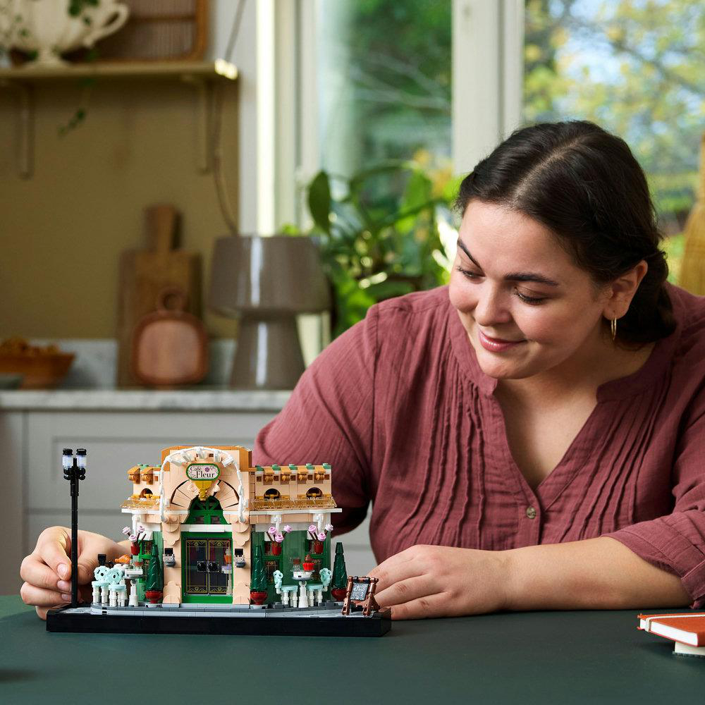 Lächelnde Frau, die ein LEGO-Blumenladenmodell auf einem dunklen Tisch mit neutralem Hintergrund hält.