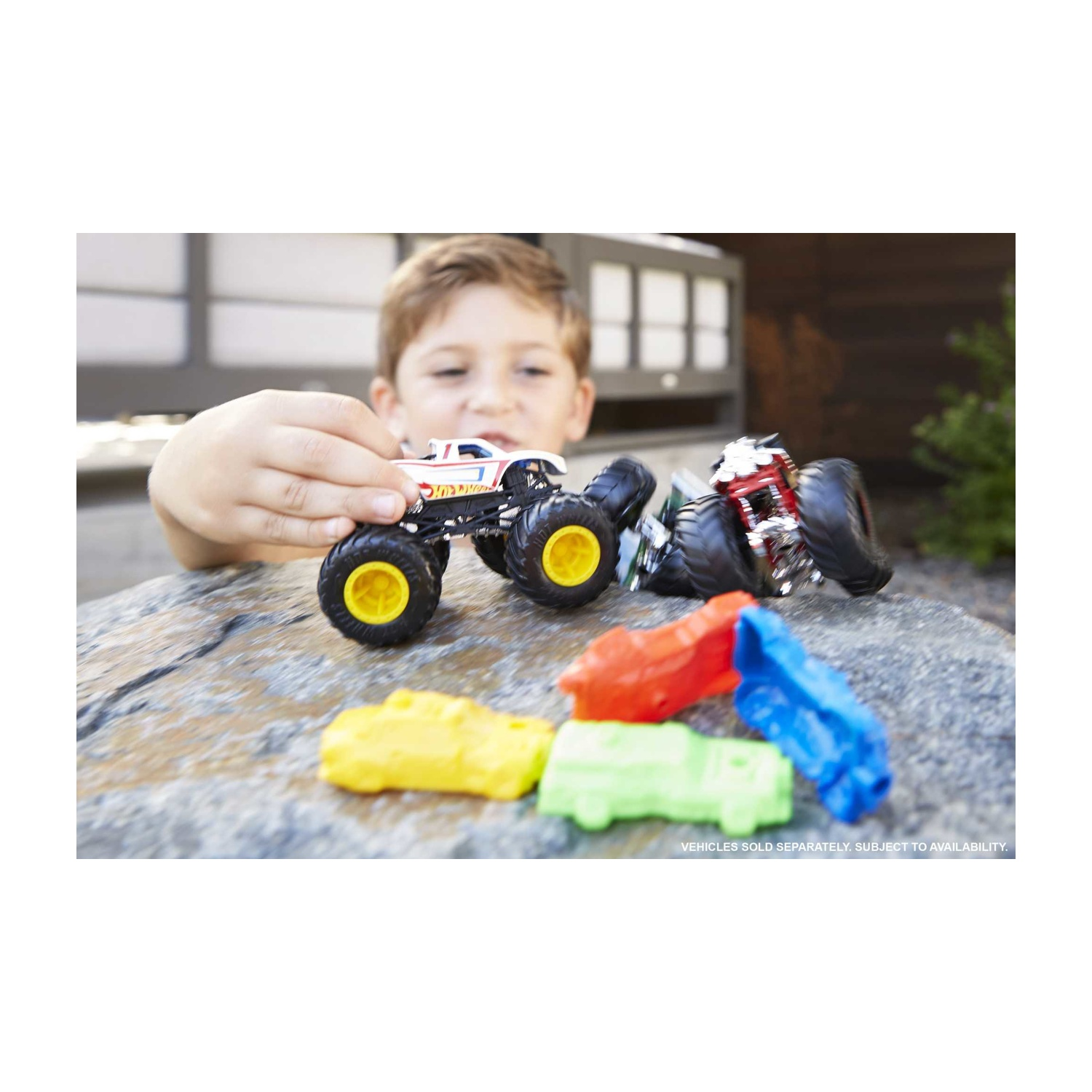 Un bambino gioca con due monster truck. Ci sono macchinine per terra. Lo sfondo è sfocato.