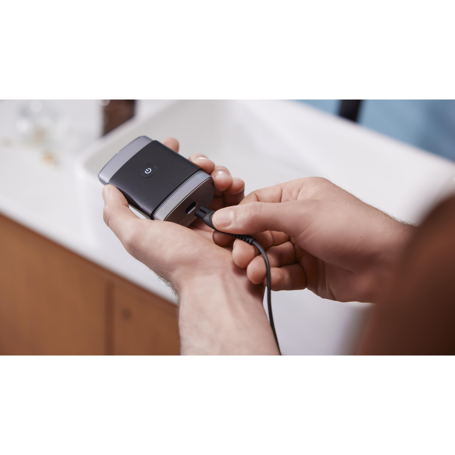 Hands holding black electric shaver and charging cable, in bathroom.
