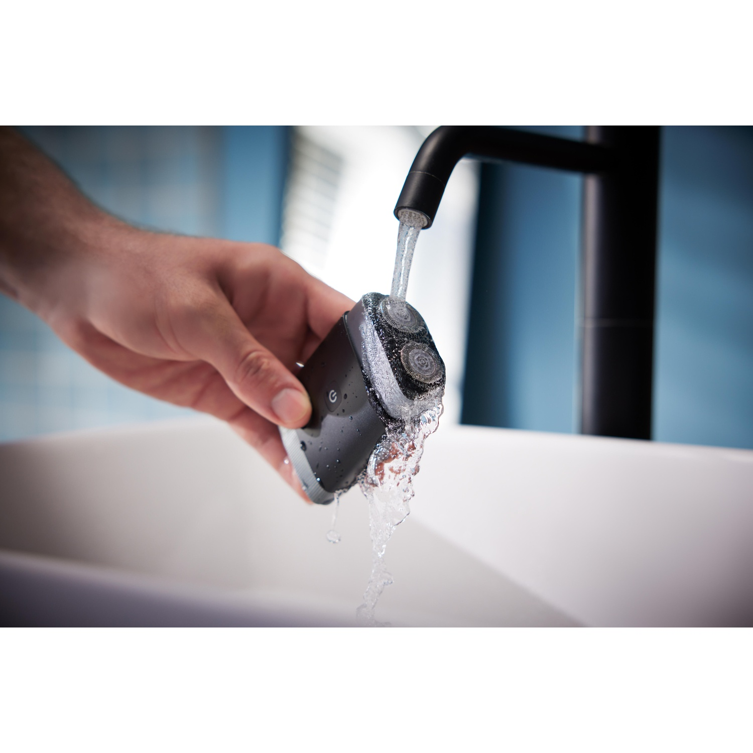 A dark gray electric shaver being rinsed under running water in sink.