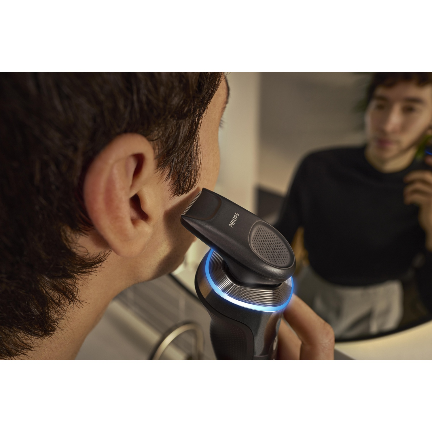 Man shaves his face with a black and silver Philips electric shaver in a mirror.