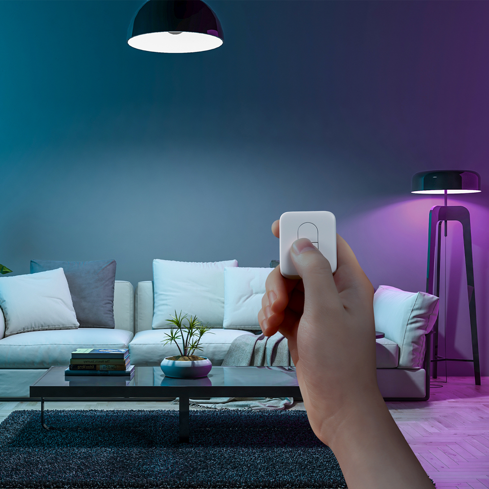 Hand, die eine weiße Fernbedienung in einem Raum hält und die Lichter steuert. Couch, Lampe und Pflanze sind im Hintergrund.