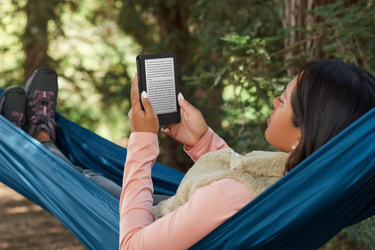 Nő függőágyban Kindle e-olvasót olvas elmosódott zöld fák előtt.