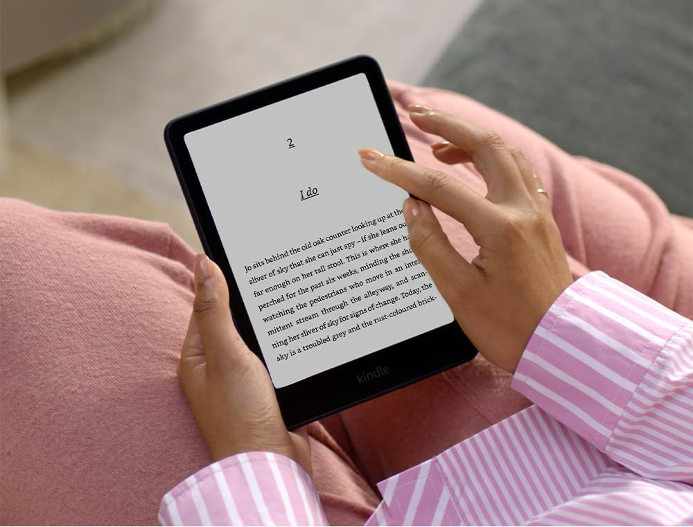 Kéz egy fekete Kindle-t tart. Az olvasó egy könyvoldalon van szöveggel. A személy ül.