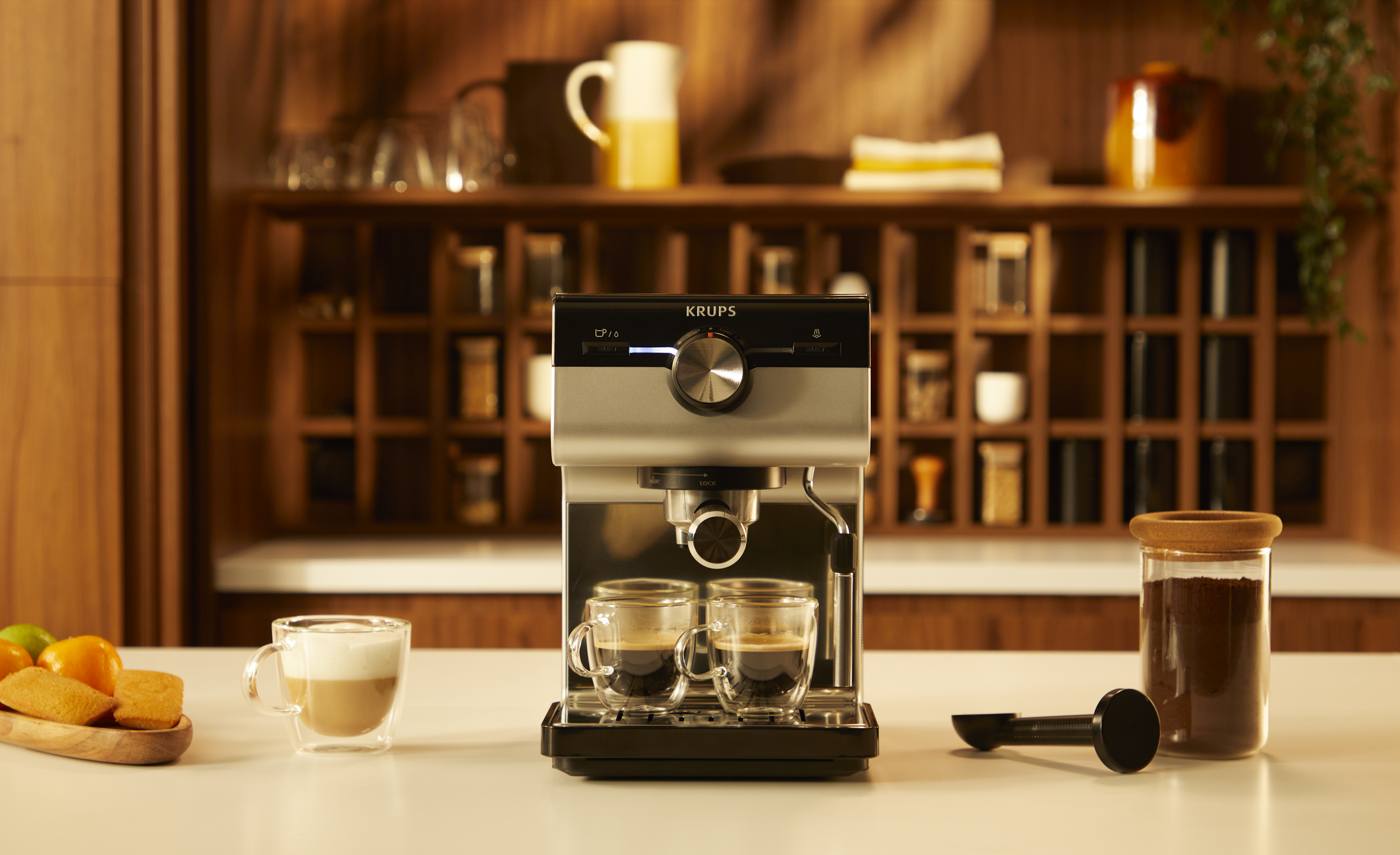 Srebrno-czarny ekspres do kawy Krups z dwiema filiżankami kawy na białej powierzchni.