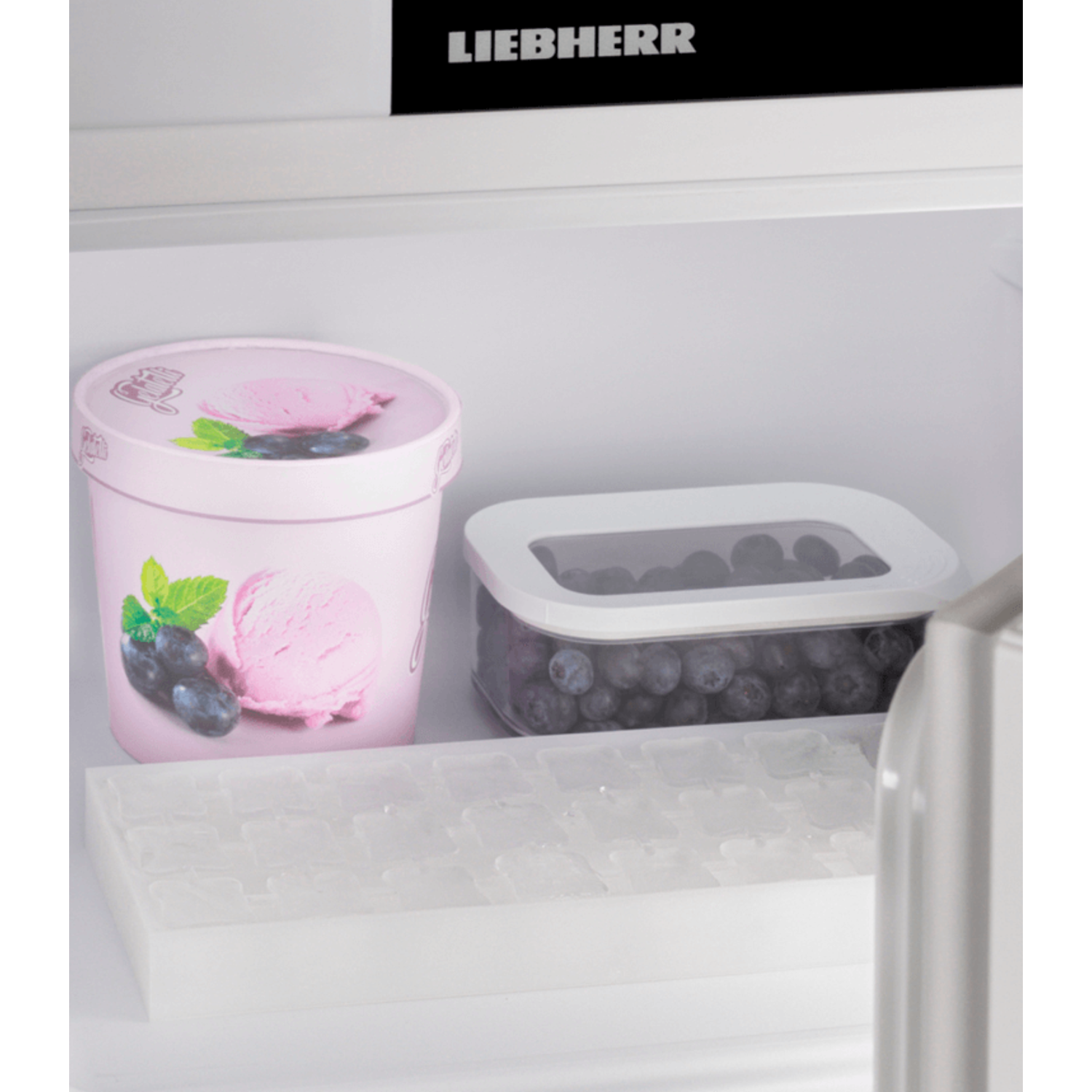 Fridge interior with ice tray, ice cream, and blueberries in a container.