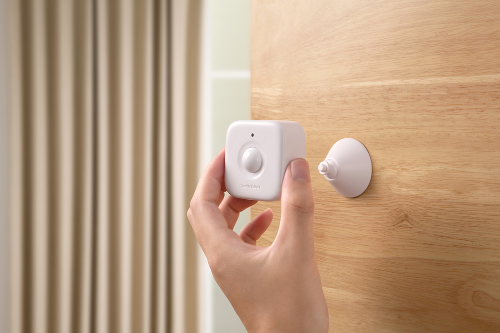 Hand hält einen weißen Smart-Home-Sensor an einer Holztür neben einem Saugnapf.