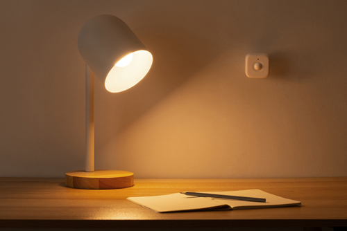 Eine beleuchtete weiße Lampe auf einem Holzschreibtisch mit Notizbuch und Stift. Ein Wandgerät ist sichtbar.