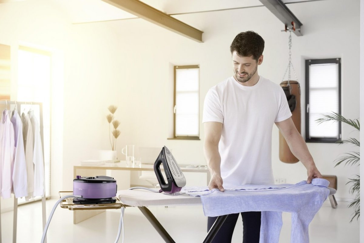 Mężczyzna w białej koszulce prasuje niebieską koszulę na desce do prasowania w dobrze oświetlonym pokoju.