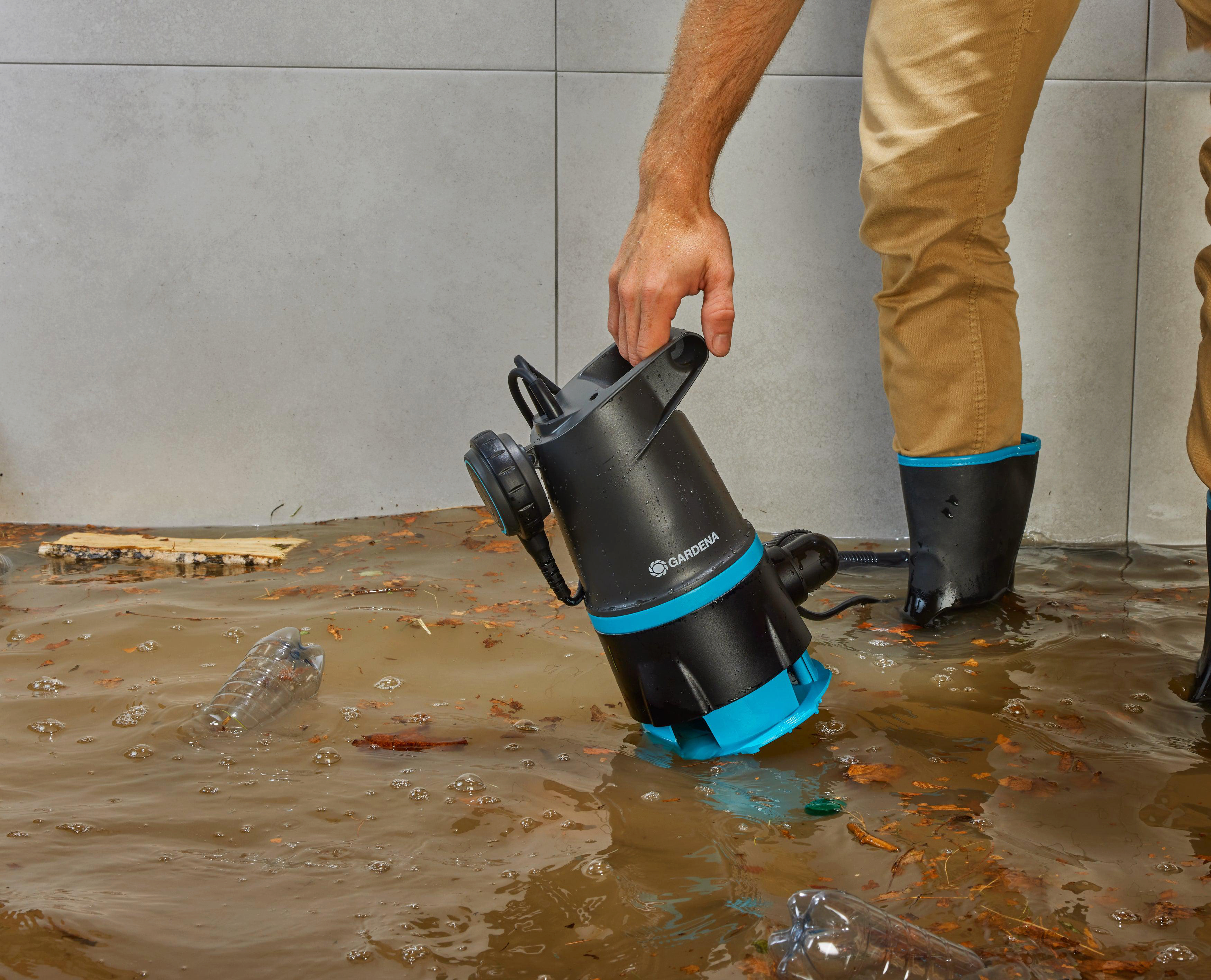 Eine Person hält eine schwarz-blaue Wasserpumpe in überflutetem braunem Wasser mit Trümmern.