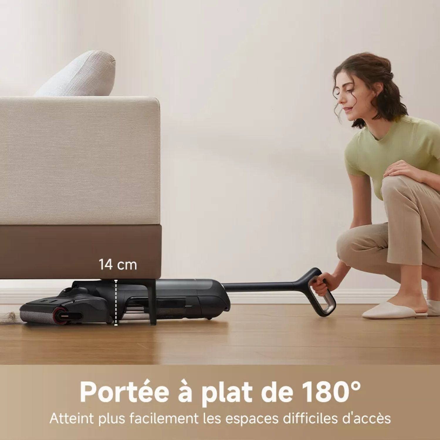 Une femme passe l'aspirateur sous un canapé. L'aspirateur est noir et a une longue poignée.