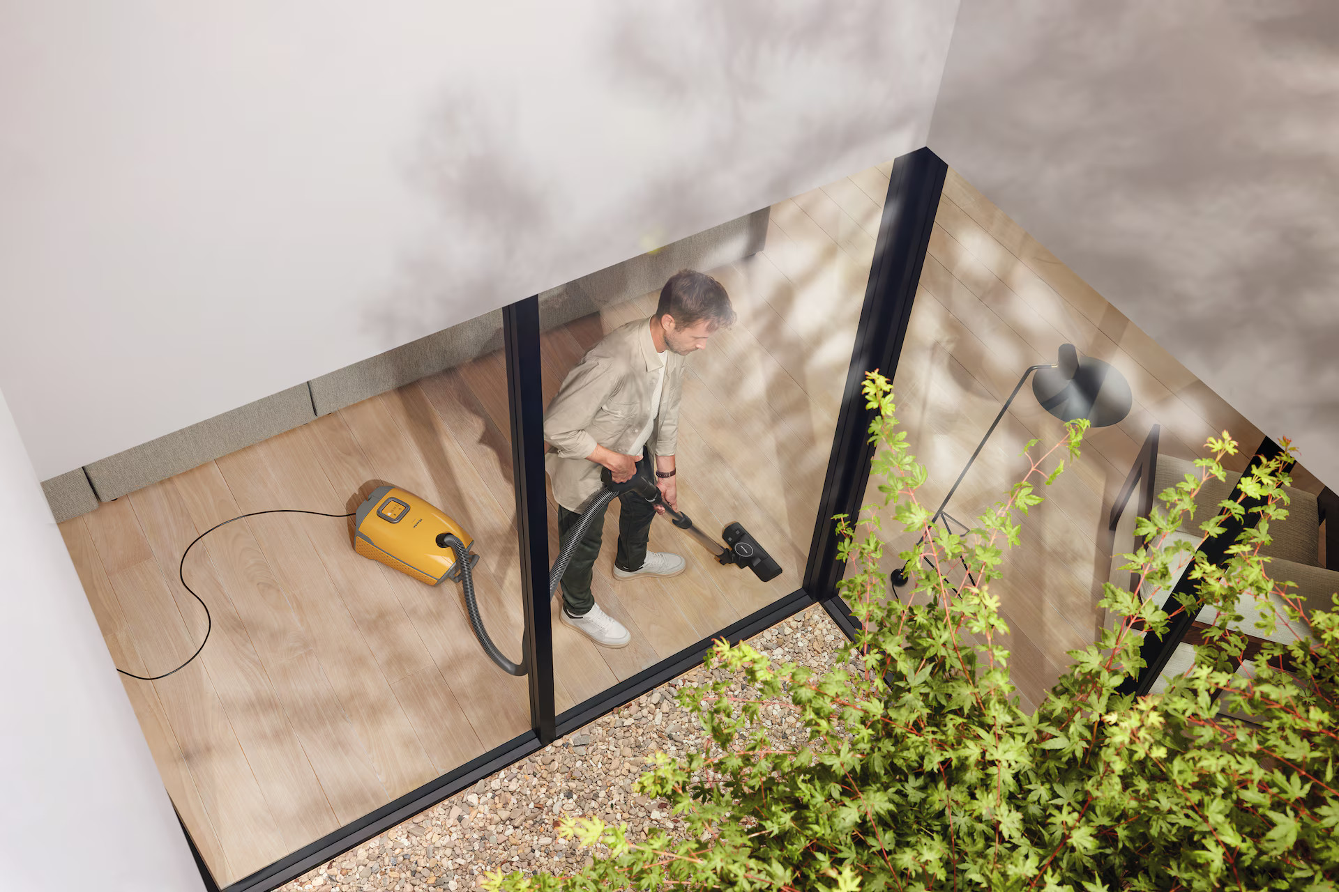 Mężczyzna odkurza drewnianą podłogę. Odkurzacz jest żółty. Duże okno jest widoczne po prawej stronie.