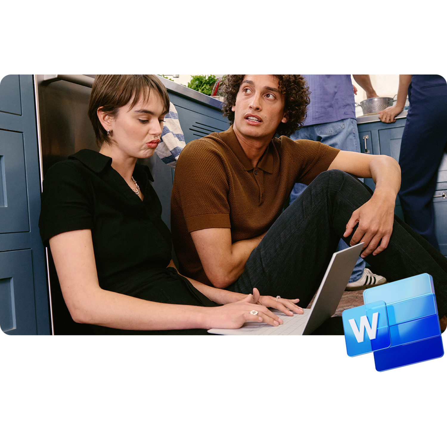 A woman and a man looking at a laptop, seated in a kitchen with others in the background.