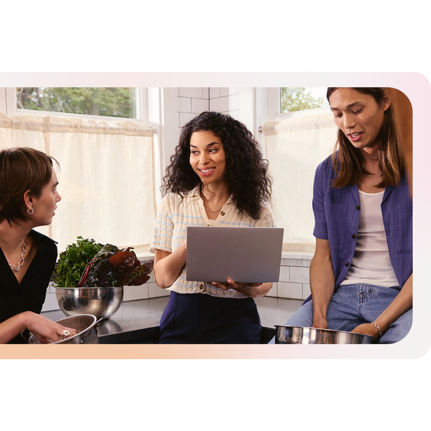 Three people in a kitchen, one with a laptop. Vegetables and metal bowls are on the counter.