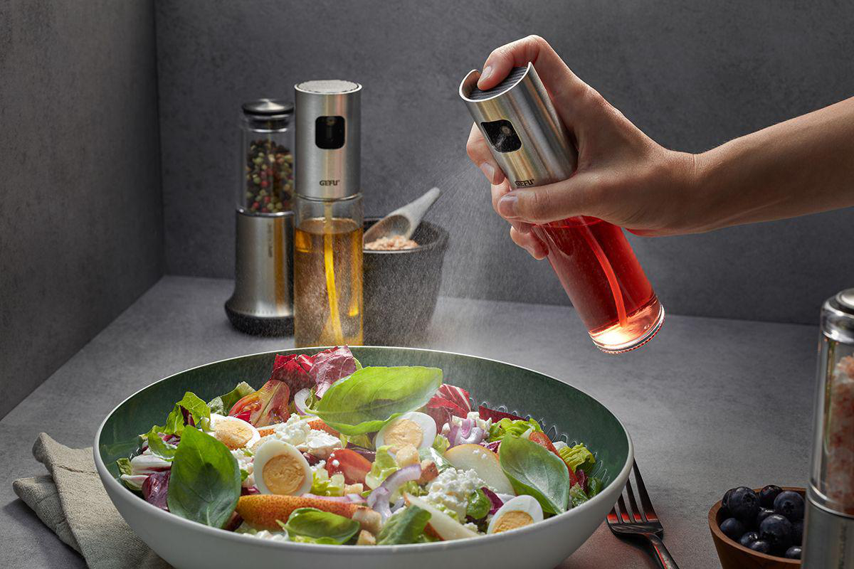 Person sprüht rotes Öl über einen Salat. Salat in grüner Schüssel, andere Ölflaschen im Hintergrund.