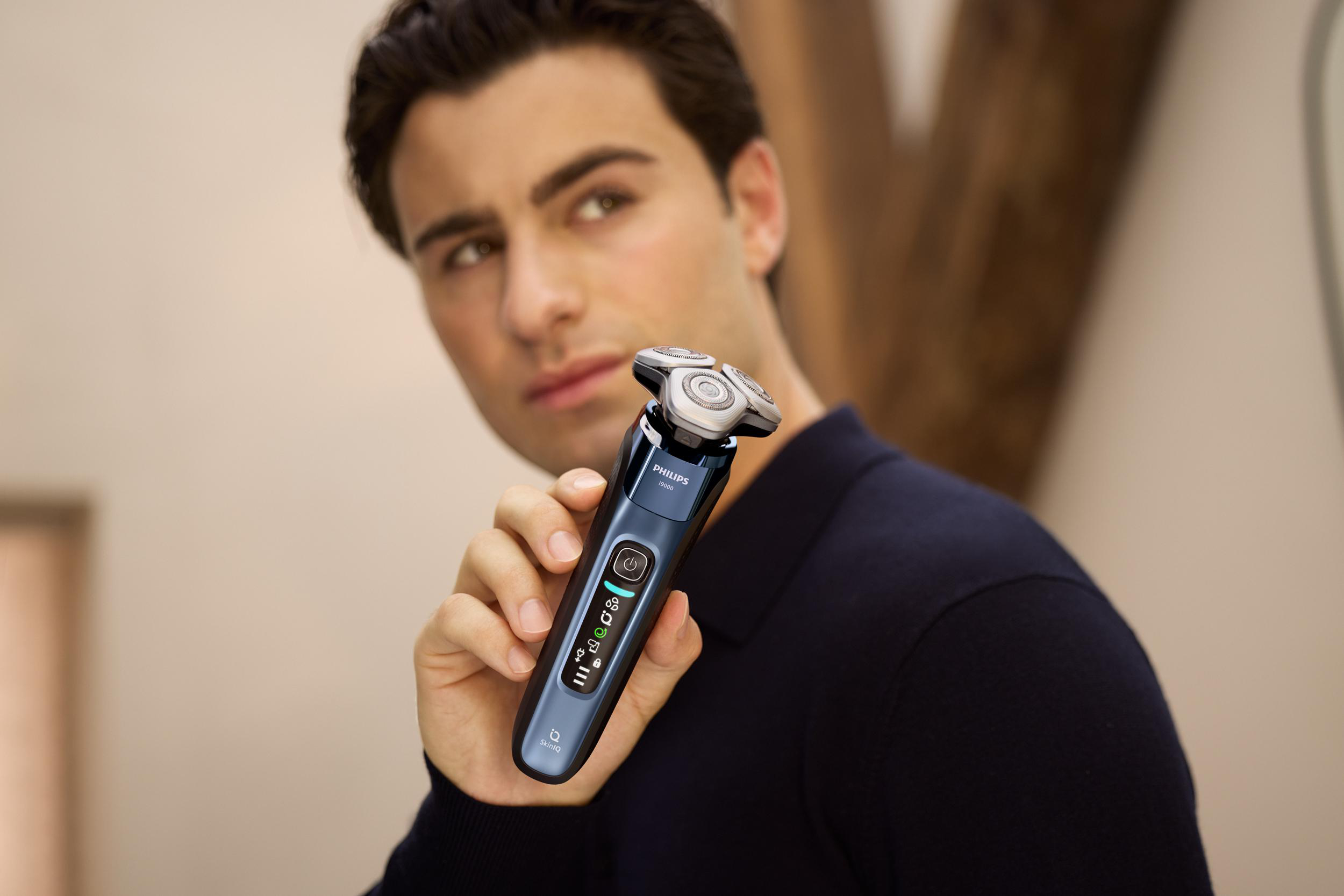 Mann hält einen blau-schwarzen Elektrorasierer. Im Hintergrund Spiegel und Holzbalken.