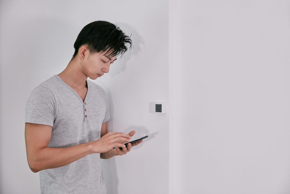 Mann in grauem Hemd betrachtet Smartphone nahe weißer Wand und quadratischem Gerät.
