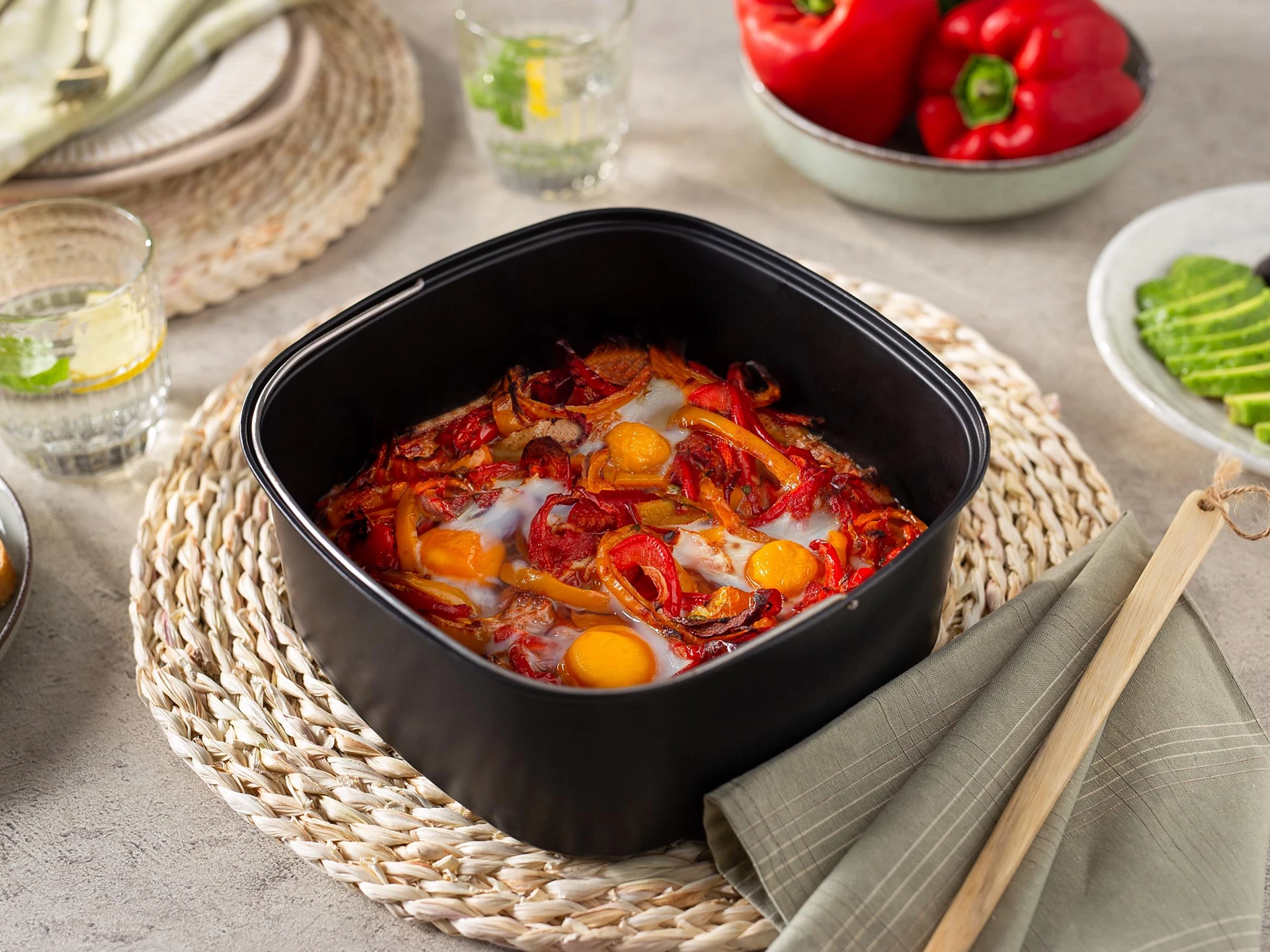 Eine Heißluftfritteuse enthält Eier, Paprika und Tomaten. Sie steht auf einer gewebten Matte mit Beilagen.