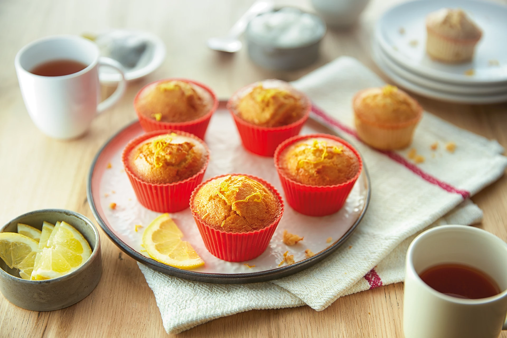 Muffins in roten Papierförmchen auf einem rosa Teller, serviert mit Tee und Zitronen.
