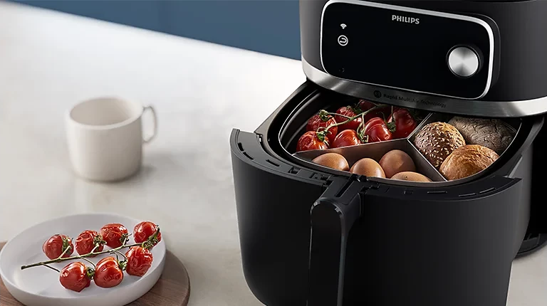Schwarze Heißluftfritteuse mit Essen darin und einem Teller Tomaten davor. Weiße Tasse daneben.