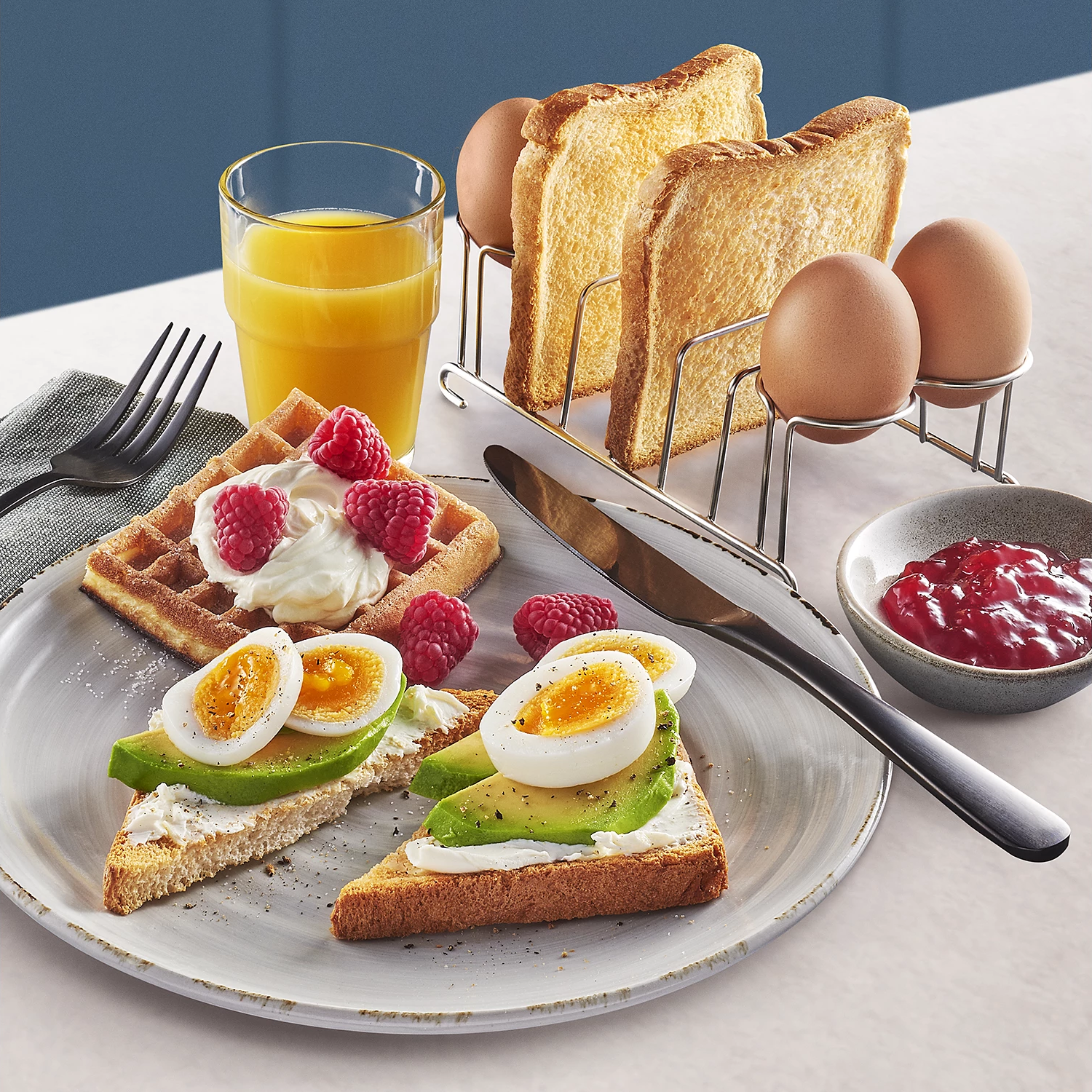Eine Frühstücksszene mit Waffeln, Toast, Eiern und einem Glas Orangensaft. Enthält Marmelade und ein Messer.
