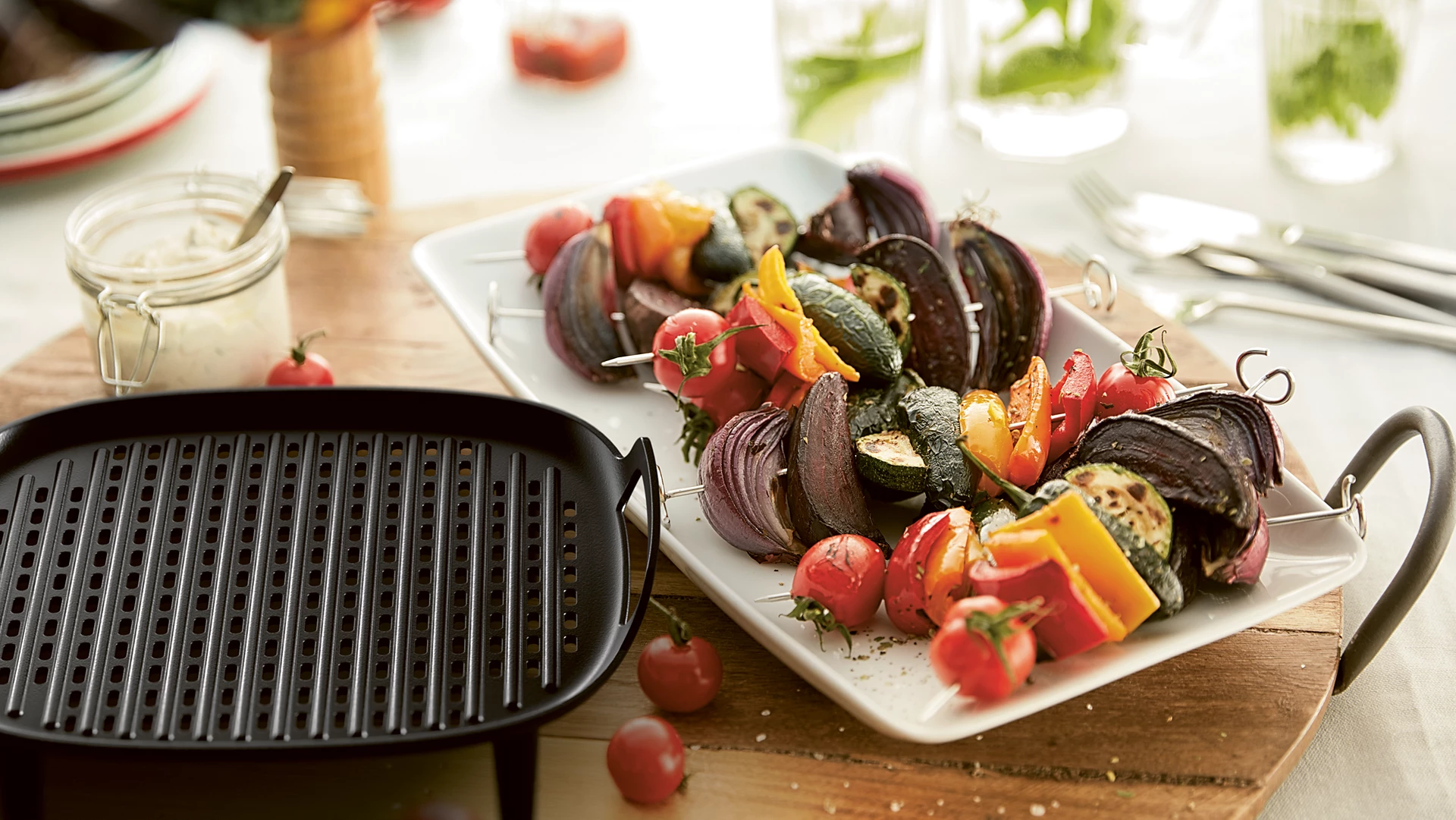Gegrilltes Gemüse und Fleischspieße auf weißem Tablett, mit Grillpfanne und Soße.