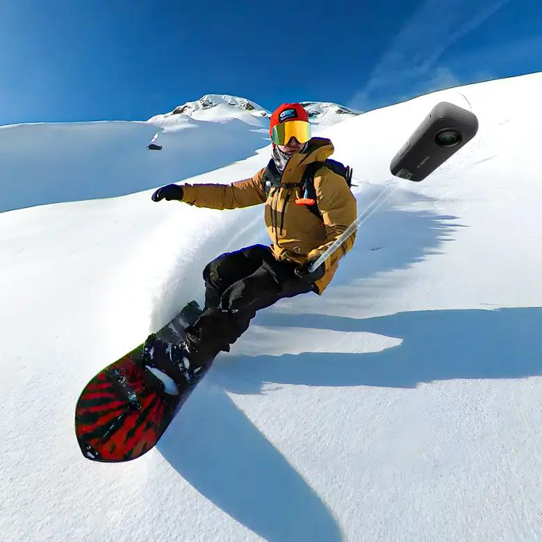 Snowboarder auf verschneiter Piste, trägt Brille und gelbe Jacke. Eine Kamera auf einem Selfie-Stick filmt die Action.
