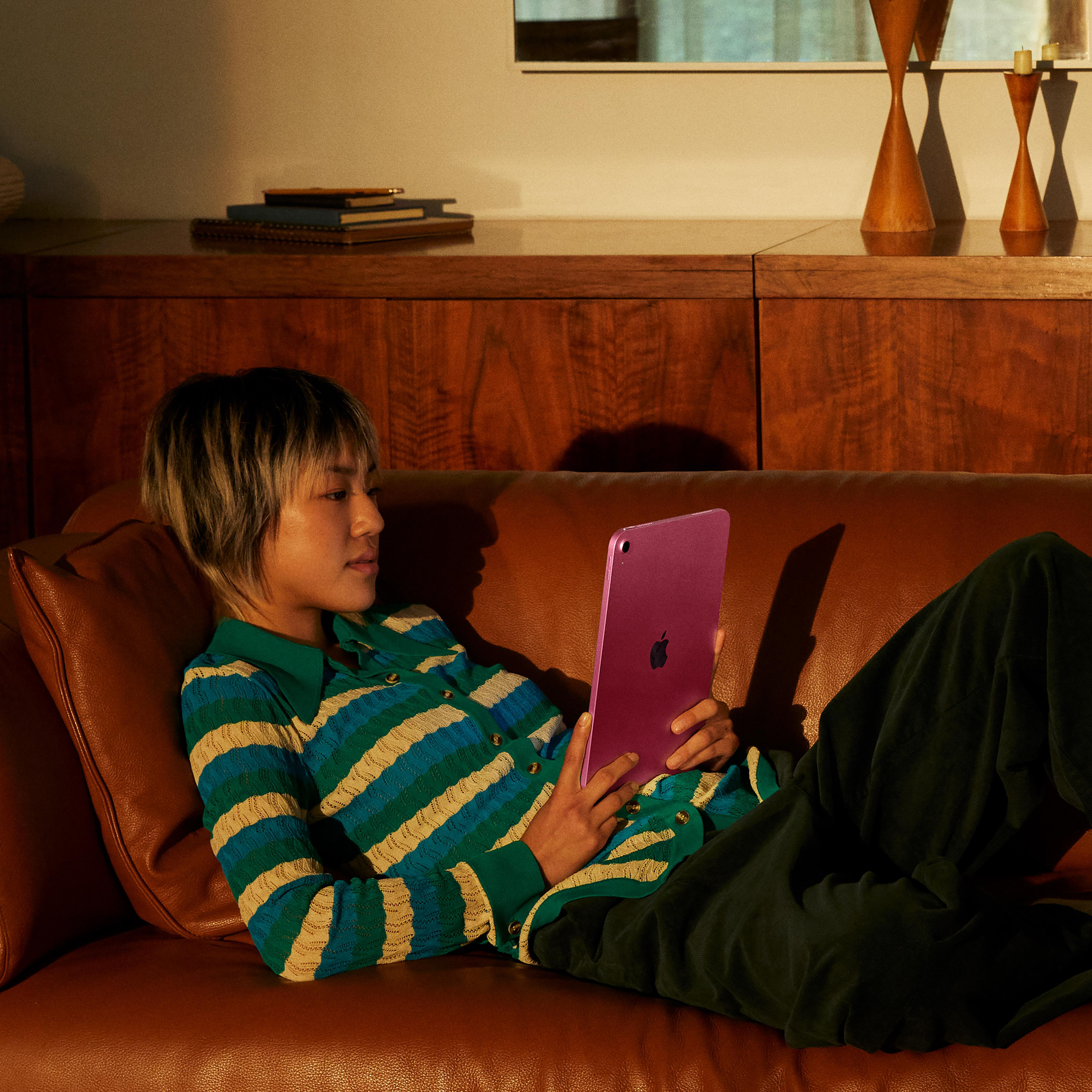 Eine Person auf einem braunen Sofa hält ein pinkfarbenes Tablet. Sie trägt ein gestreiftes Hemd und eine dunkelgrüne Hose. Holzdekor im Hintergrund.